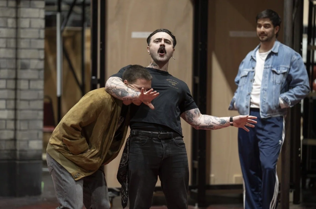 Three men in a casual setting, two of them engaged in a comedic interaction while the third looks on amused. One man is leaning over, covering his face with his hand, the second man appears to be singing or shouting, with tattoos visible on his arms, and the third man stands nearby smiling, dressed in a denim jacket and track pants.