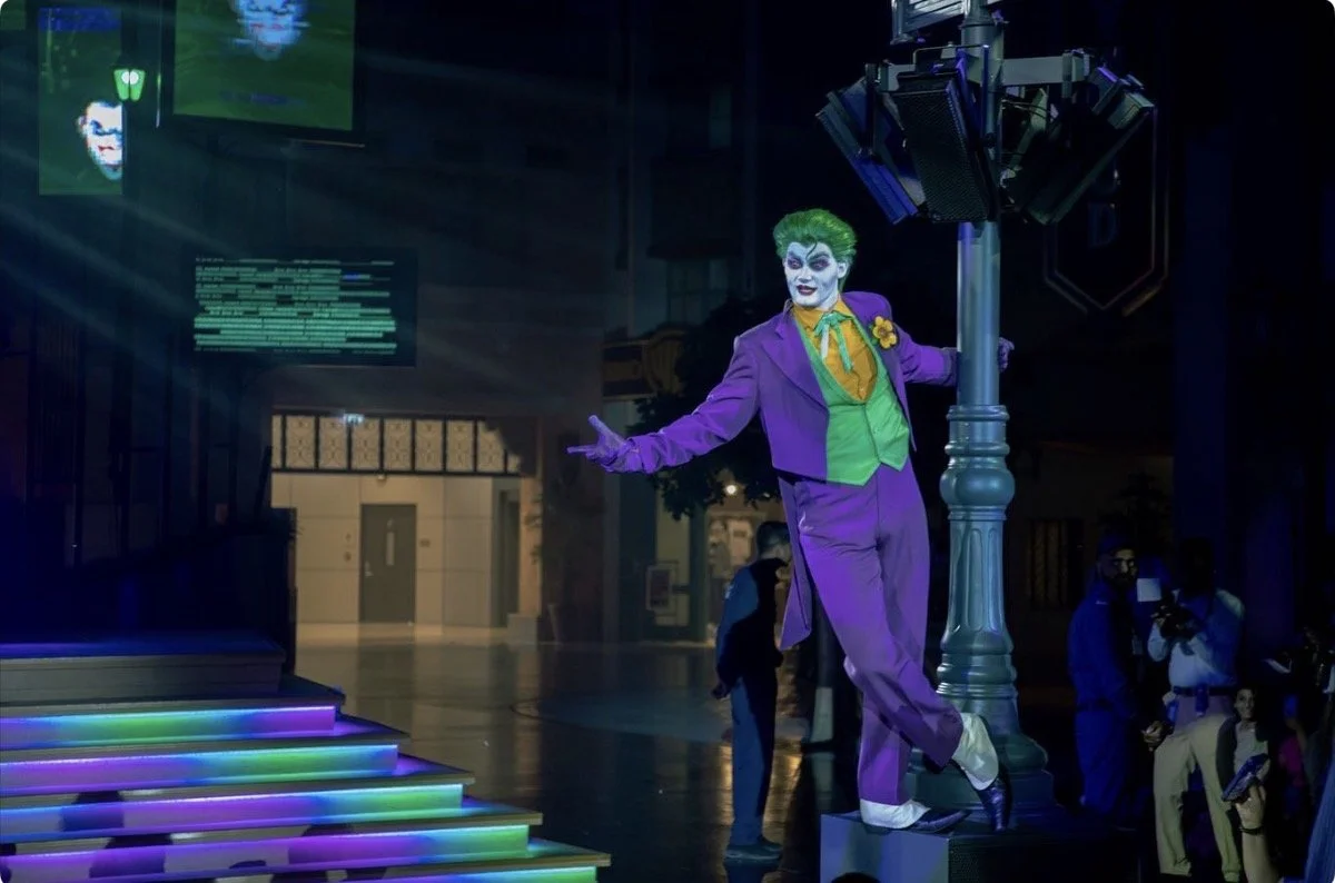 The Joker, a clown with green hair and white face paint, wearing a purple suit with a green vest and yellow shirt, standing on a lit platform and posing with arms outstretched in a dark indoor setting.
