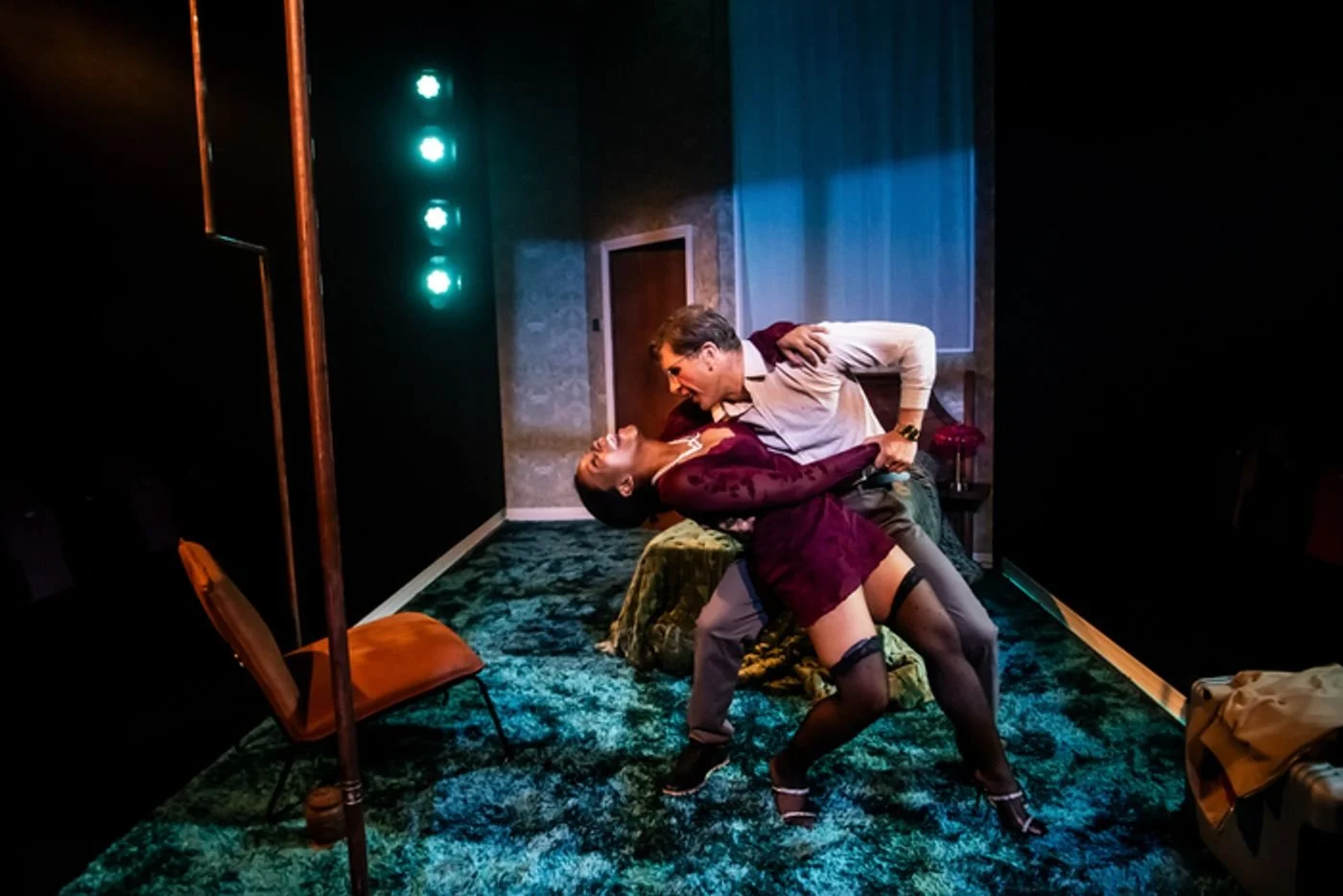 A man and woman dancing passionately in a dimly lit room with green and blue lighting. The woman is in a burgundy dress and stockings, and the man in a white shirt and gray pants. They are close, with the woman leaning back and the man leaning forward.