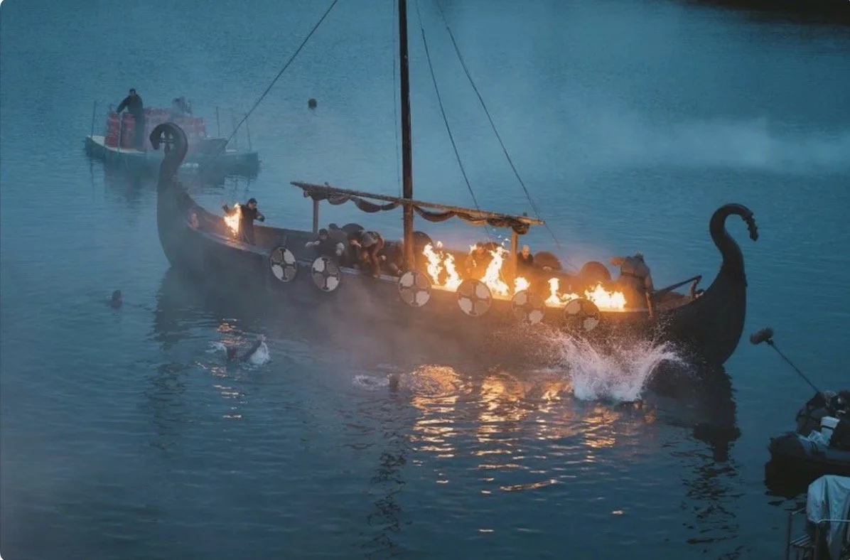 A boat with the shape of a dragon, on fire, floating on water with people on board, during dusk or nighttime.