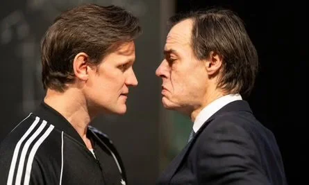 Two men face each other closely, one wearing a black jacket with white stripes and the other in a dark suit with a white shirt, in a tense confrontation.