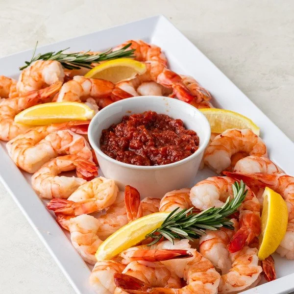 Shrimp cocktail platter with lemon wedges, rosemary sprigs, and a bowl of cocktail sauce on a white rectangular plate.