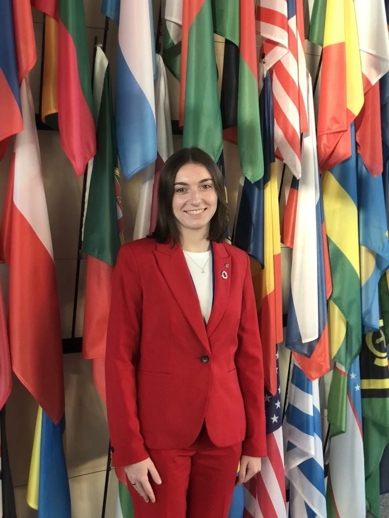 A young woman in a red suit smiling in front of a display of various national flags.