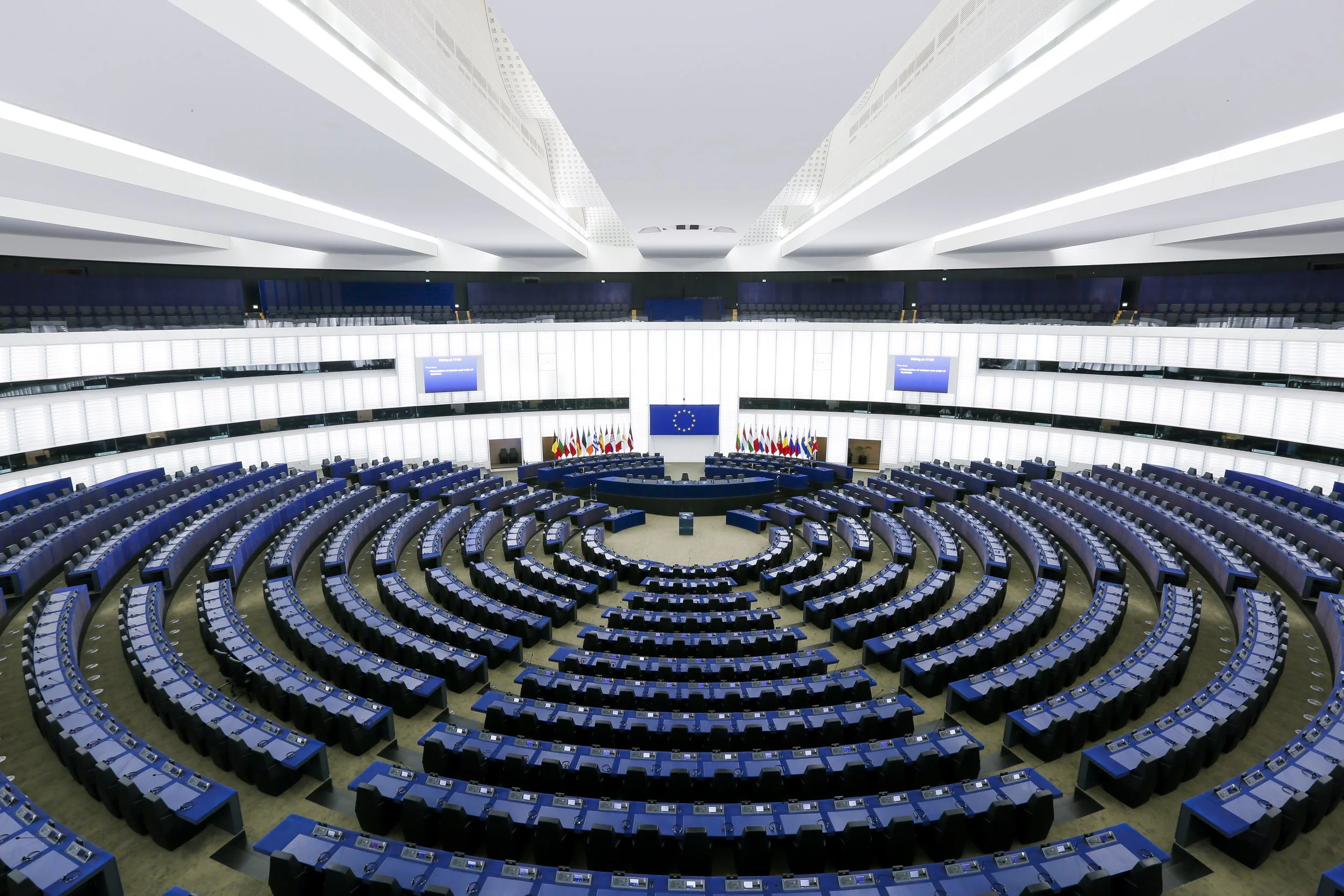Empty semicircular auditorium with blue seats arranged in concentric rows and desks, flags at the front, and multiple large screens displaying a blue background.
