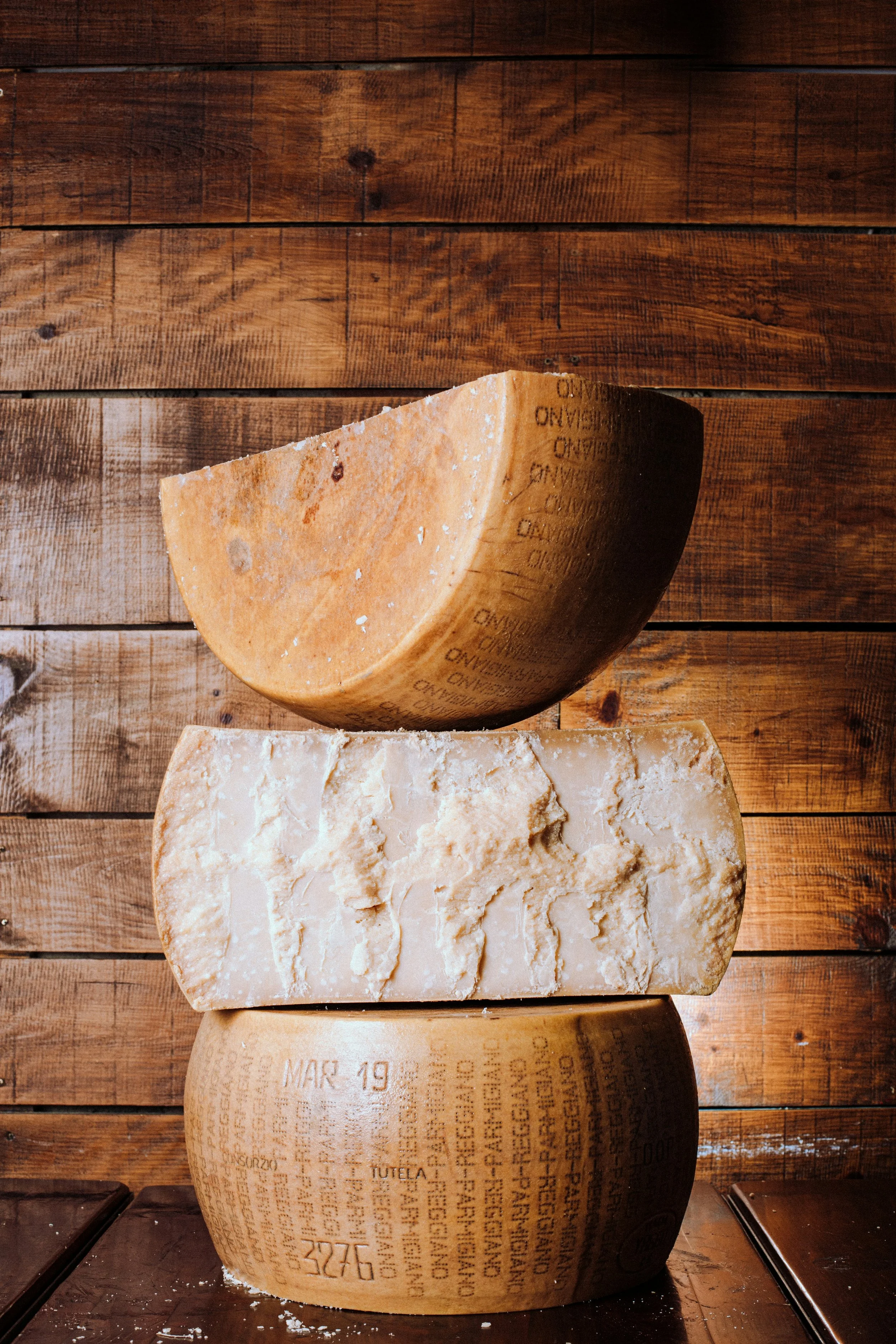 Image of three large blocks of cheese on a wooden surface. The cheeses are stacked, with a semi-hard cheese on top and a hard cheese at the bottom. The cheeses have visible textures and some text engraved on their surfaces.