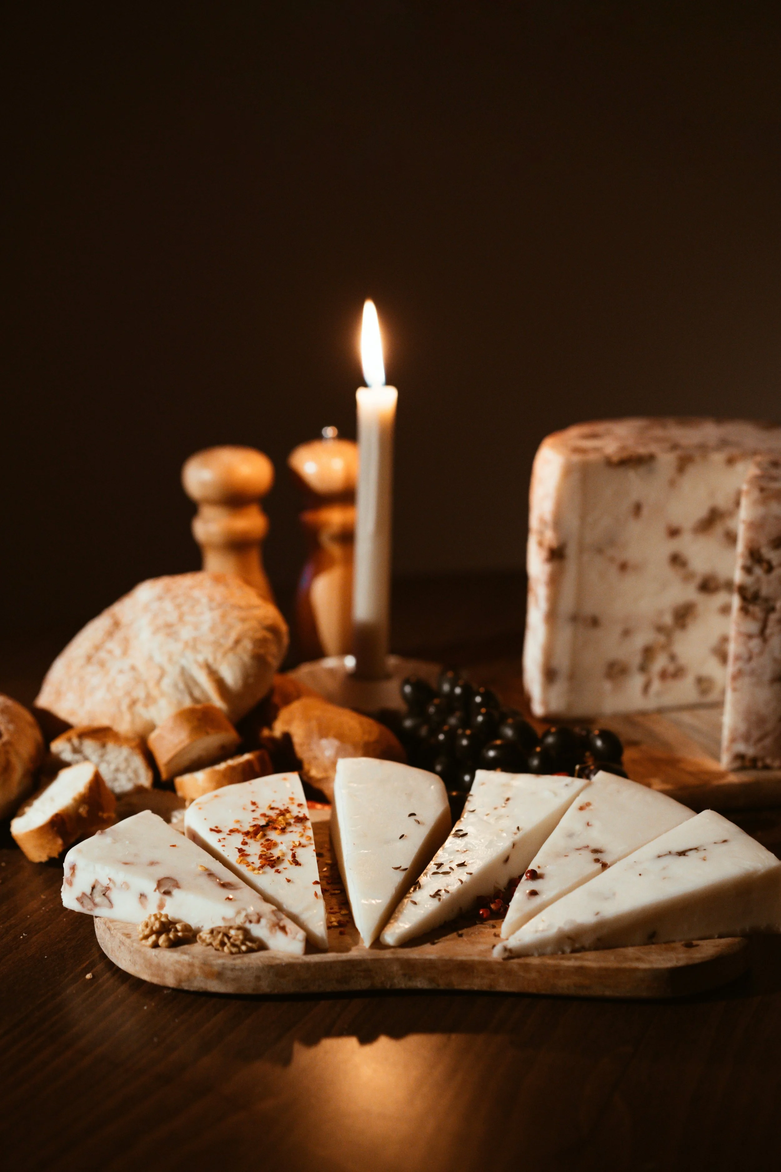 A cheese platter with sliced cheeses, bread, black olives, and a candle, set against a dark background.