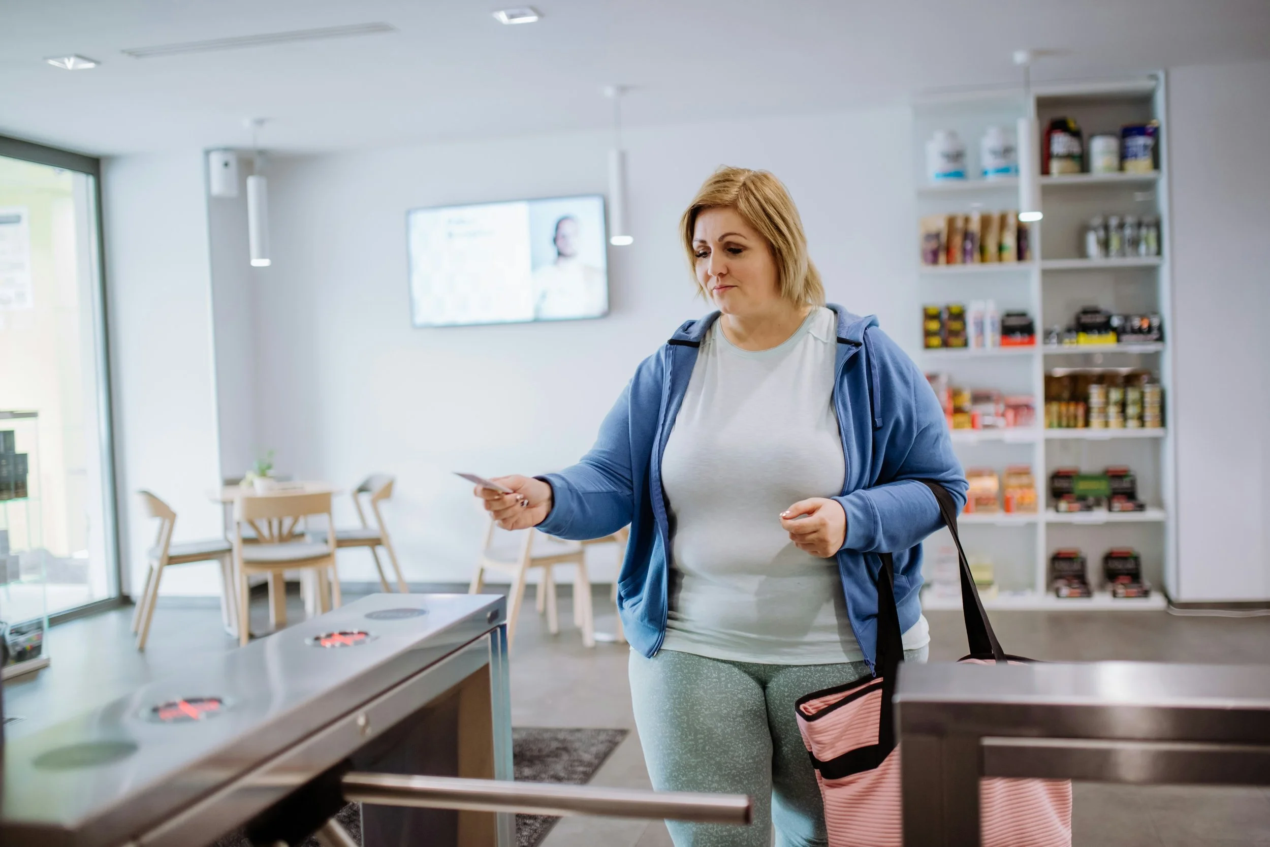 A woman with blonde hair wearing a white shirt and light green leggings shopping in a grocery store, holding a small card in her hand. The store has shelves with various products and a digital screen on the wall.