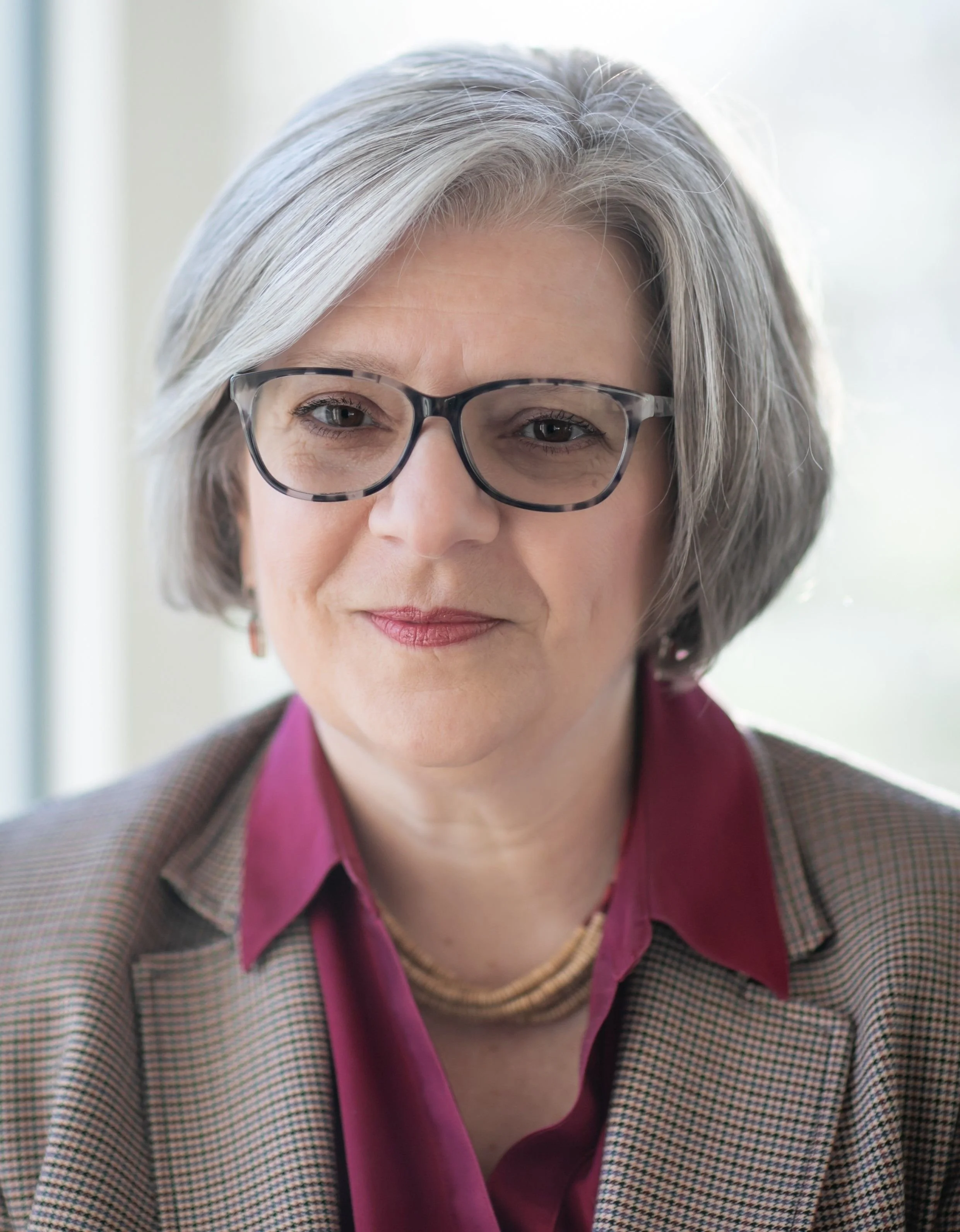 Close-up of a middle-aged woman with gray hair wearing glasses, a pink shirt, and a checkered blazer, smiling softly.