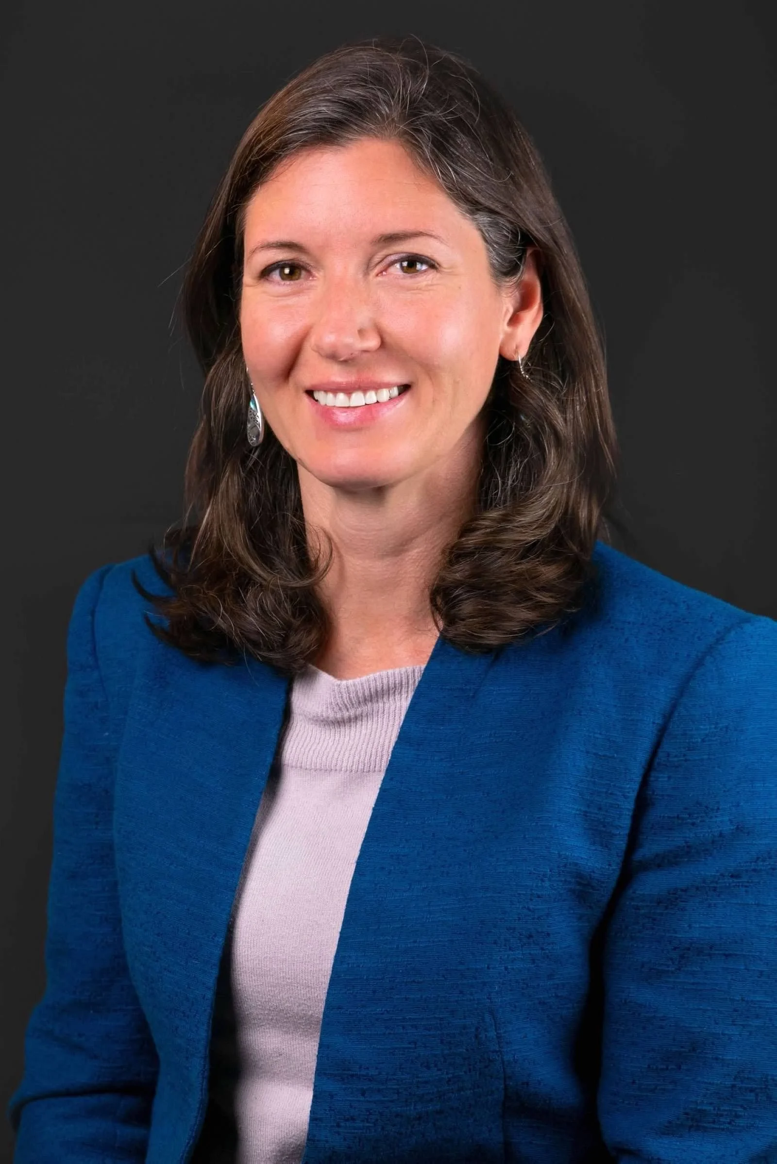 A woman with brown, shoulder-length hair wearing a blue blazer over a light pink top, smiling against a dark background.