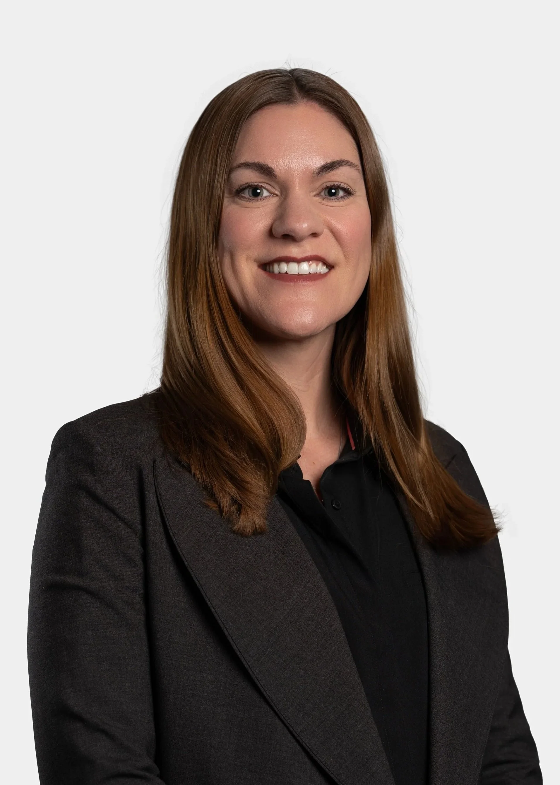 Portrait of a woman with long, wavy brown hair, wearing a black blazer and smiling against a plain white background.