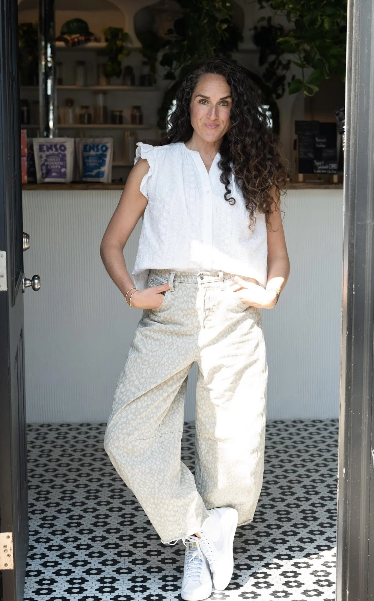 Woman with long curly hair, wearing a white sleeveless blouse, light patterned wide-leg pants, and white sneakers, standing in a doorway with hands in her pockets inside a modern cafe with geometric black and white tile floor.