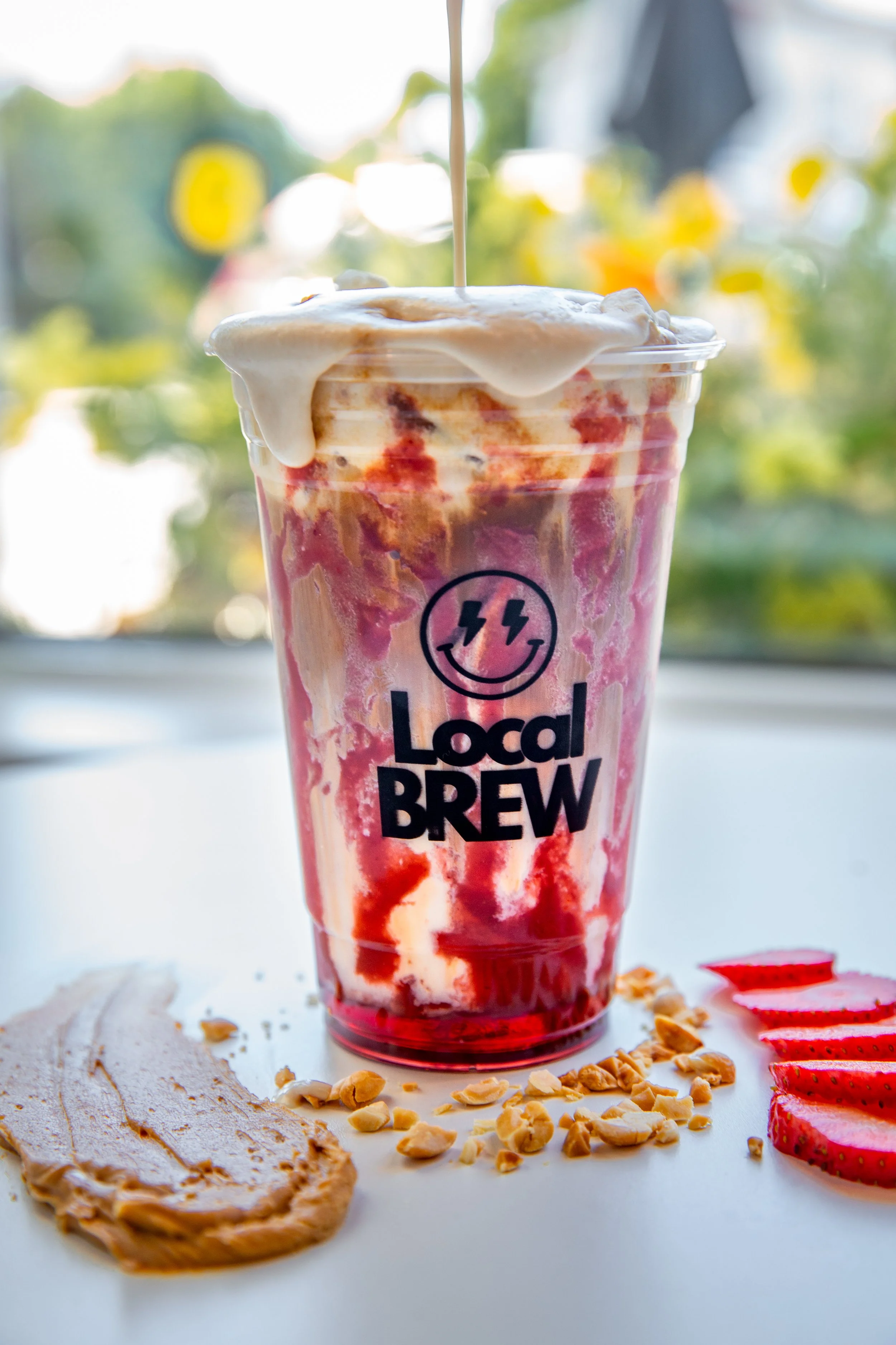 A colorful blended strawberry and cream drink in a tall plastic cup with the logo 'Local Brew' on it. The drink has whipped cream and syrup on top, with a straw inserted. Sliced strawberries, crushed nuts, and a piece of a cookie are on a white surfa