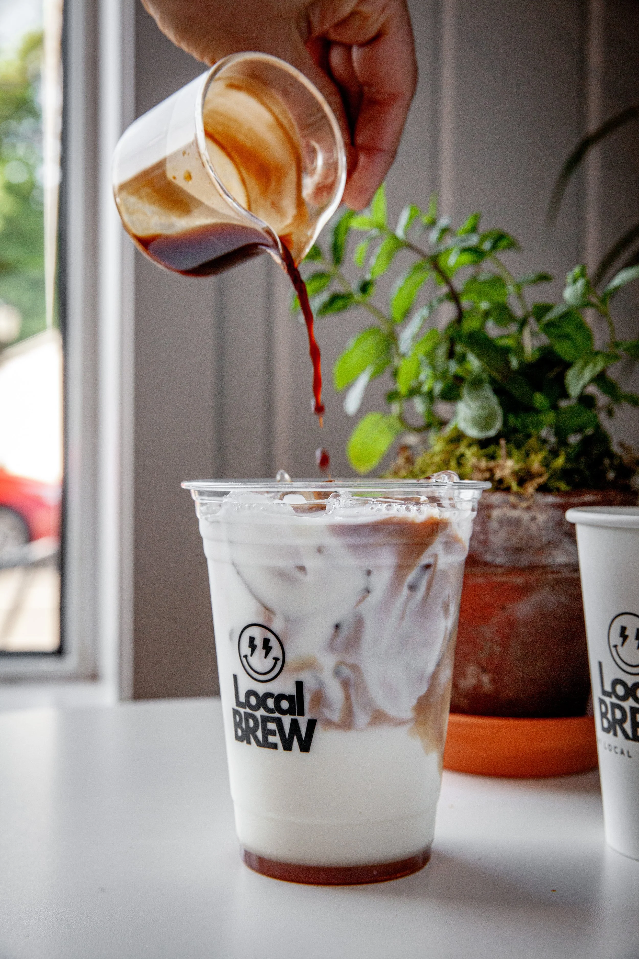 A person is pouring coffee into a cup with a 'Local Brew' logo on it, with a potted plant and window in the background.