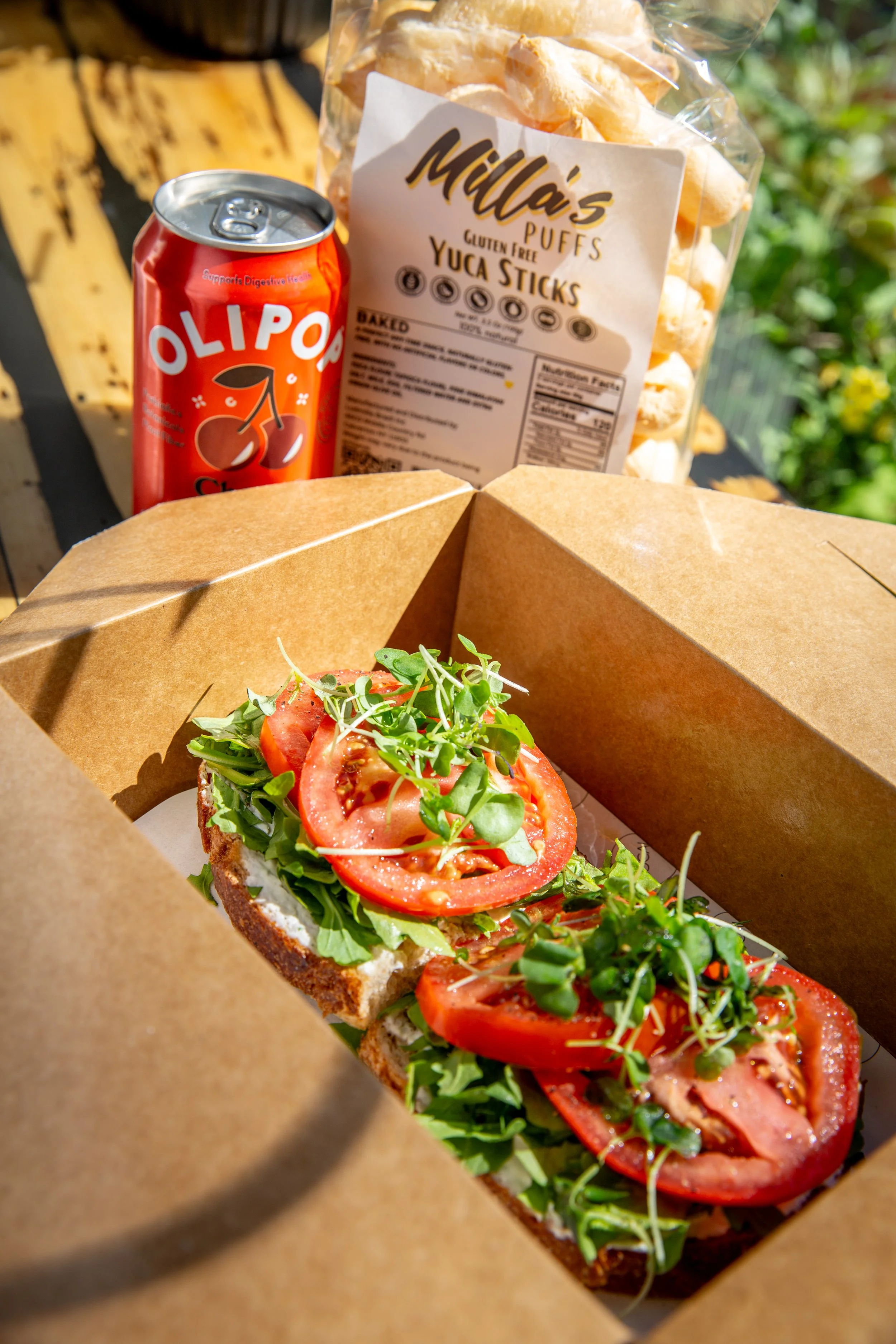 Open brown takeout box with sliced tomato, arugula, and toasted bread sandwich.