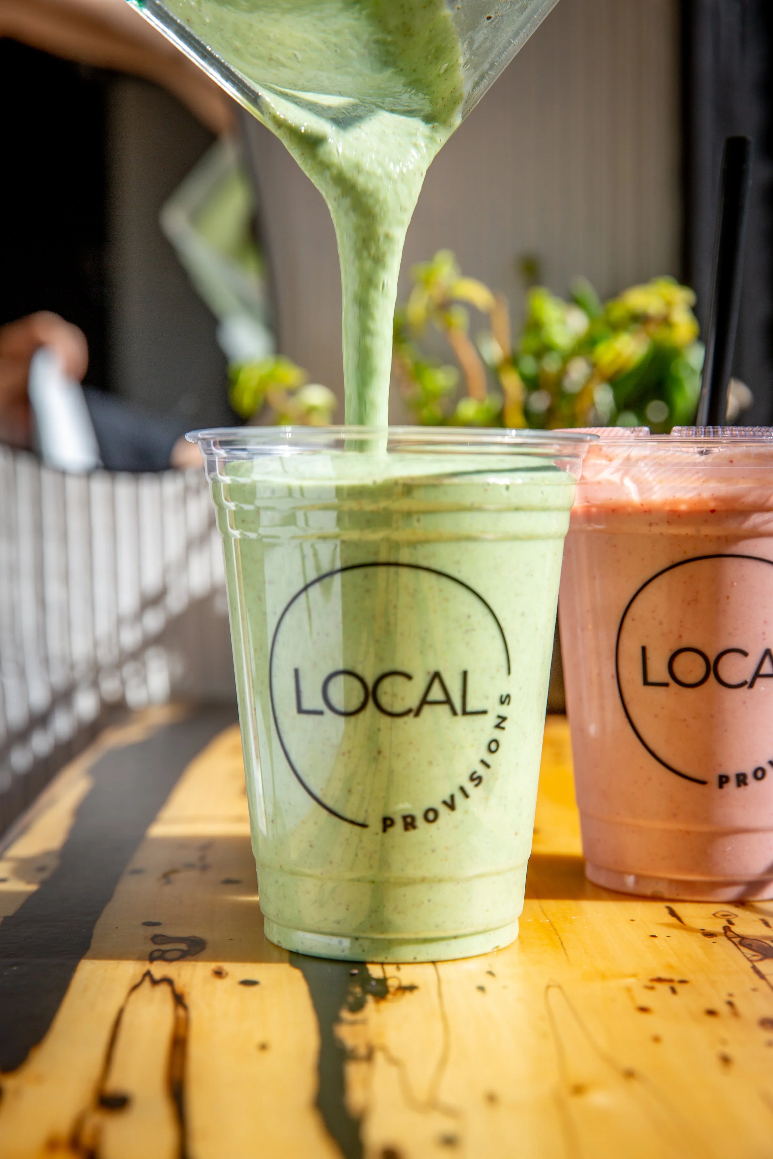 Green smoothie being poured into a clear cup with 'LOCAL' written on it, positioned beside a pink smoothie in a similar cup, both on a wooden table.