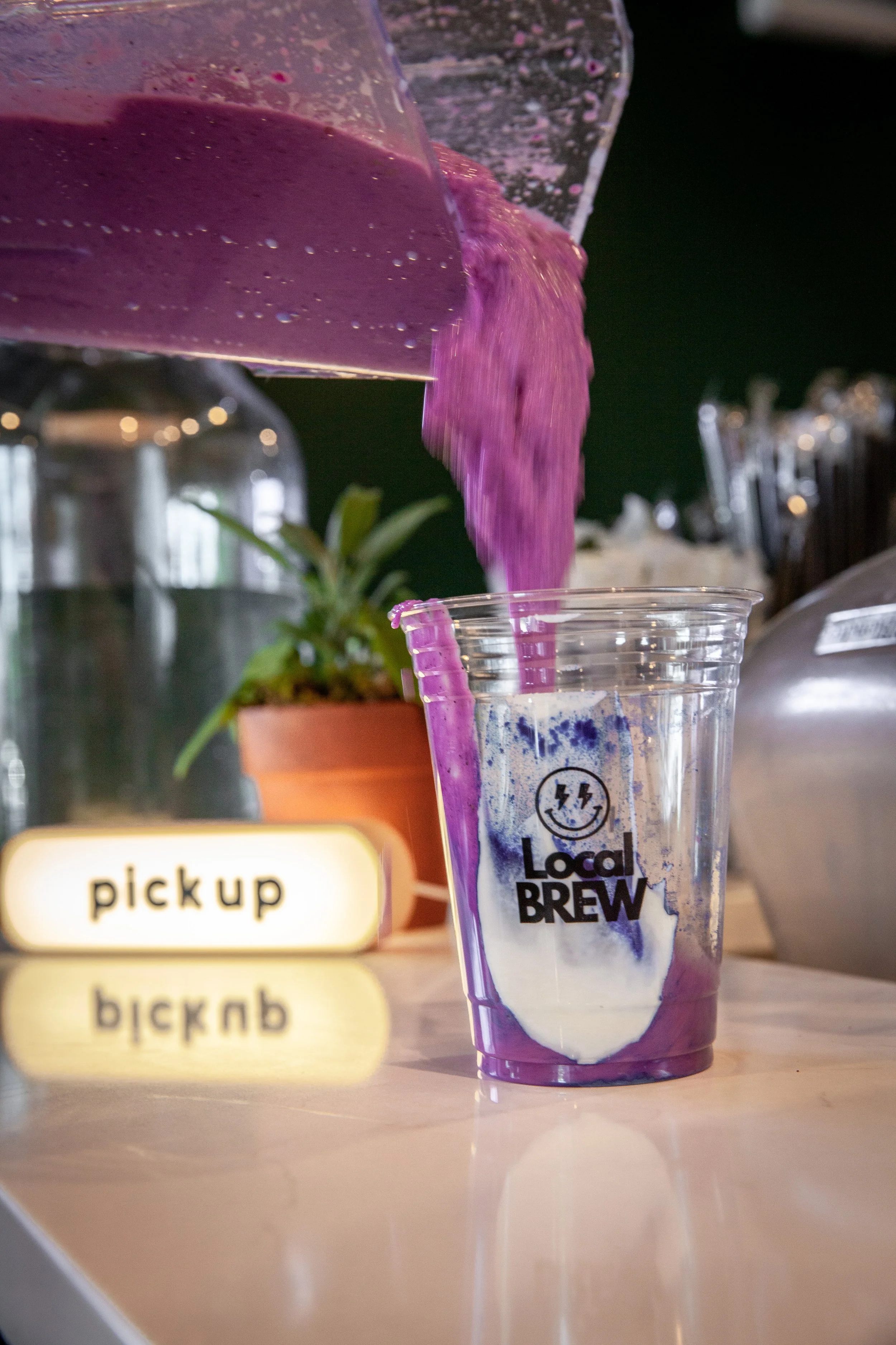 A plastic cup with purple and white swirl design and 'Local Brew' logo, filled with a purple beverage and ice, placed on a white table. Pouring purple liquid from a container into the cup, with a small potted plant and a food pickup sign visible in t