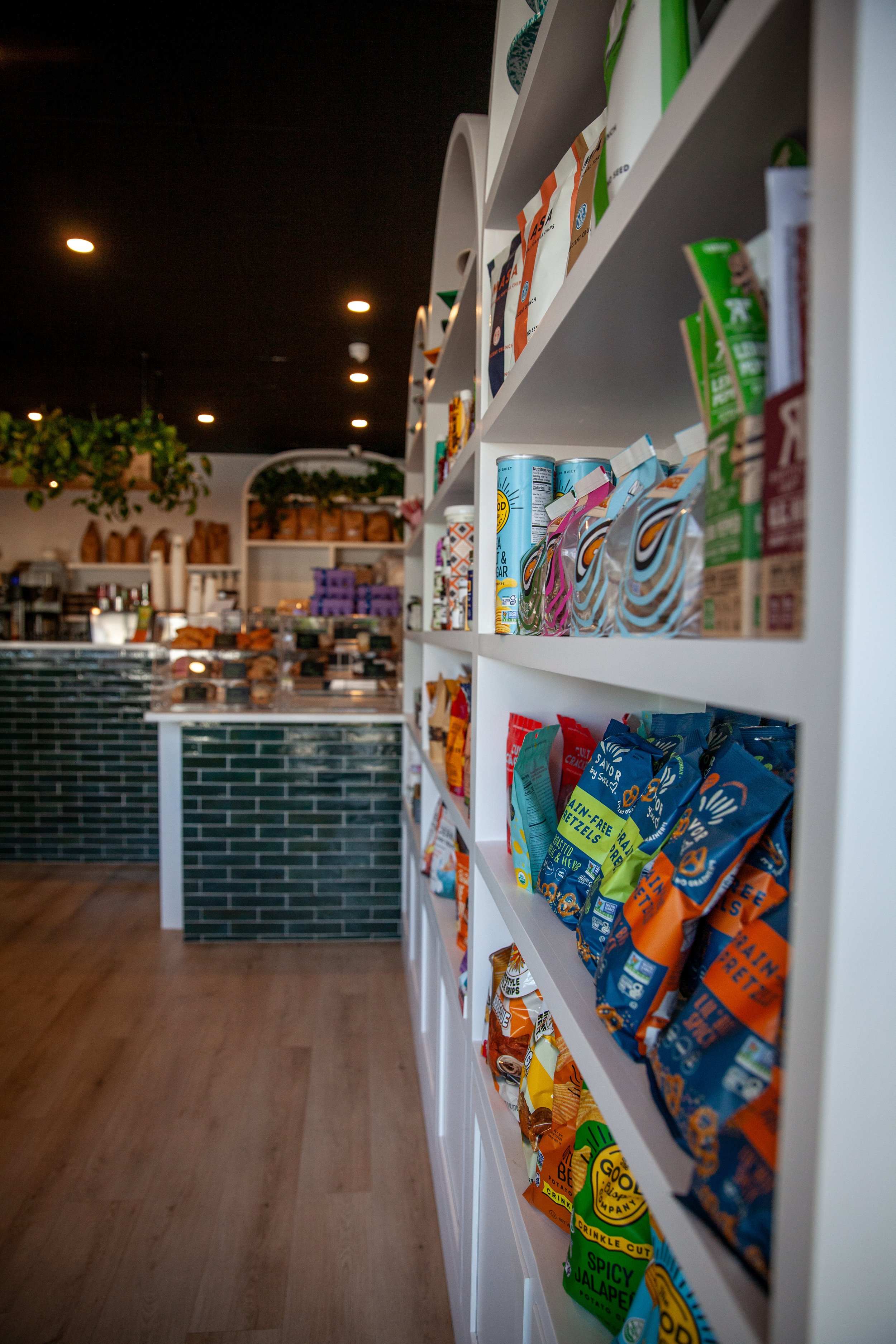 A white shelving unit stocked with various flavored potato chips in colorful bags inside a store. The store has a dark ceiling with small recessed lights and a green and black tiled counter in the background, with some greenery on top.