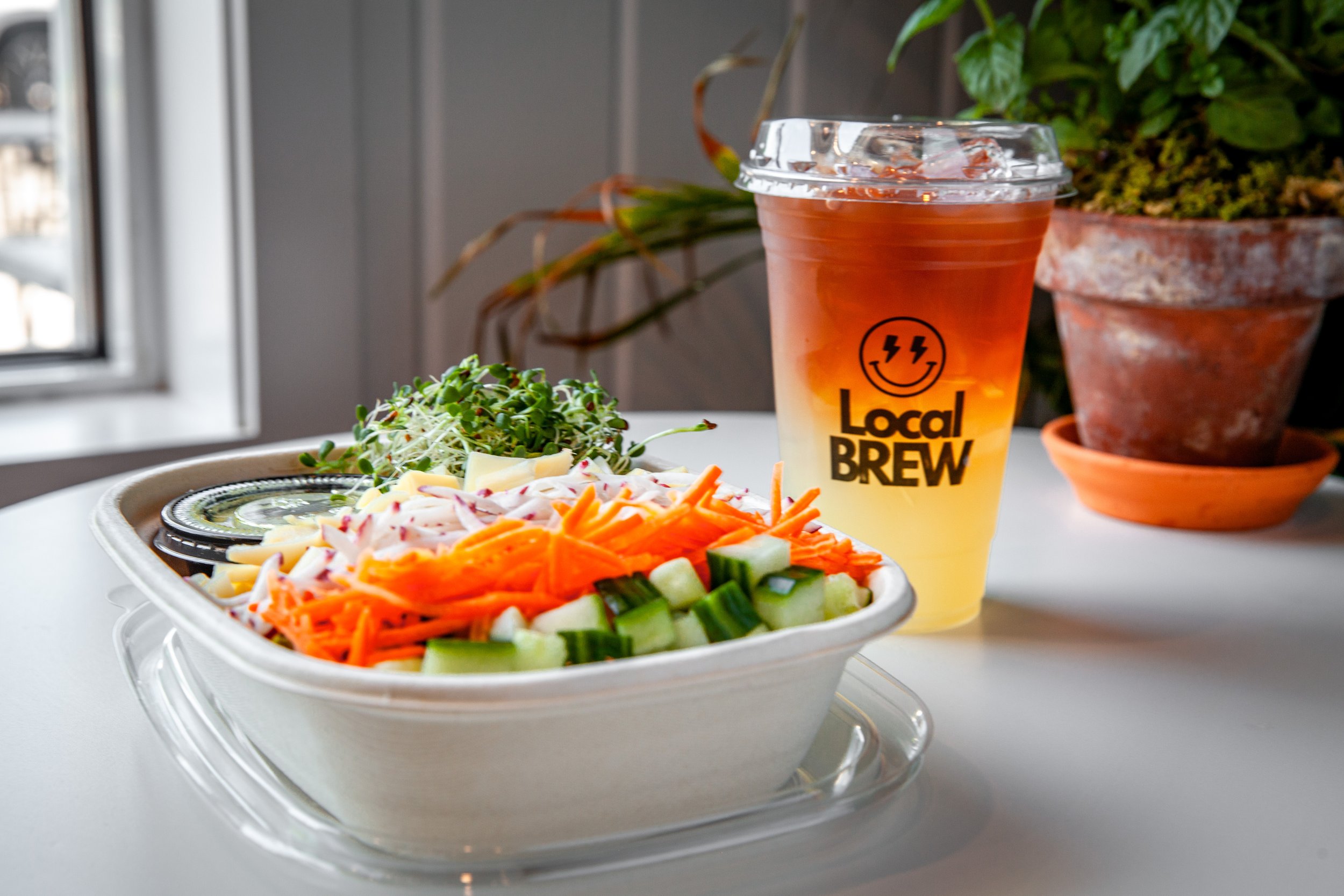Close-up of a plastic container filled with mixed shredded vegetables like carrots, cucumbers, and cabbage on a white table. In the background, there is a cup with a gradient orange and yellow beverage with a plastic lid and straw, featuring the logo