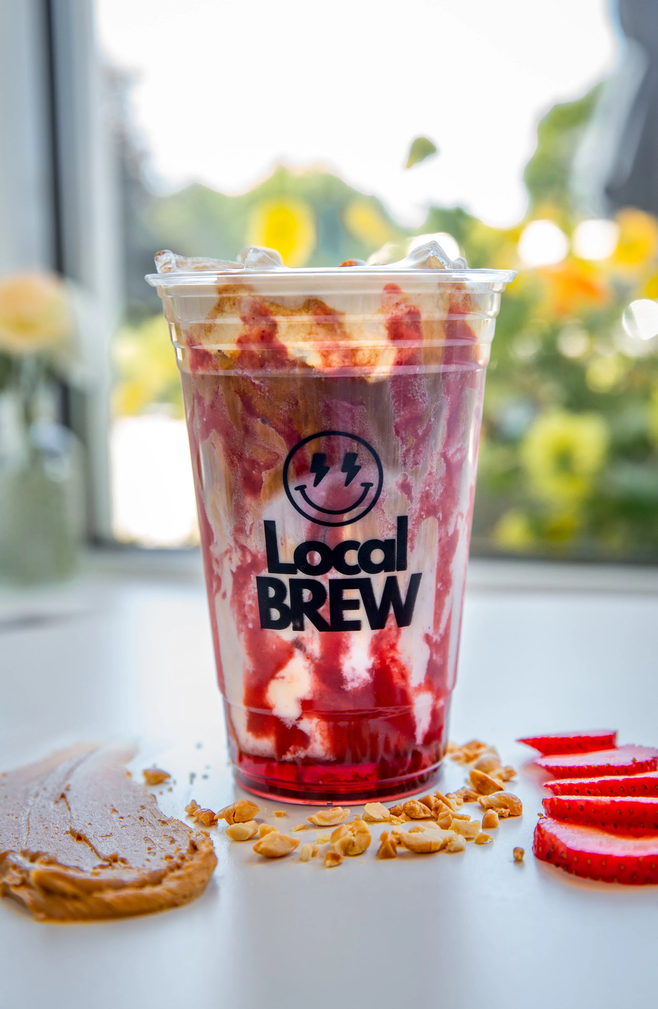 A plastic cup of red and white berry and cream smoothie from Local Brew, on a white surface with sliced strawberries, peanut butter, and scattered almond slices, with a blurred window and outdoor greenery in the background.