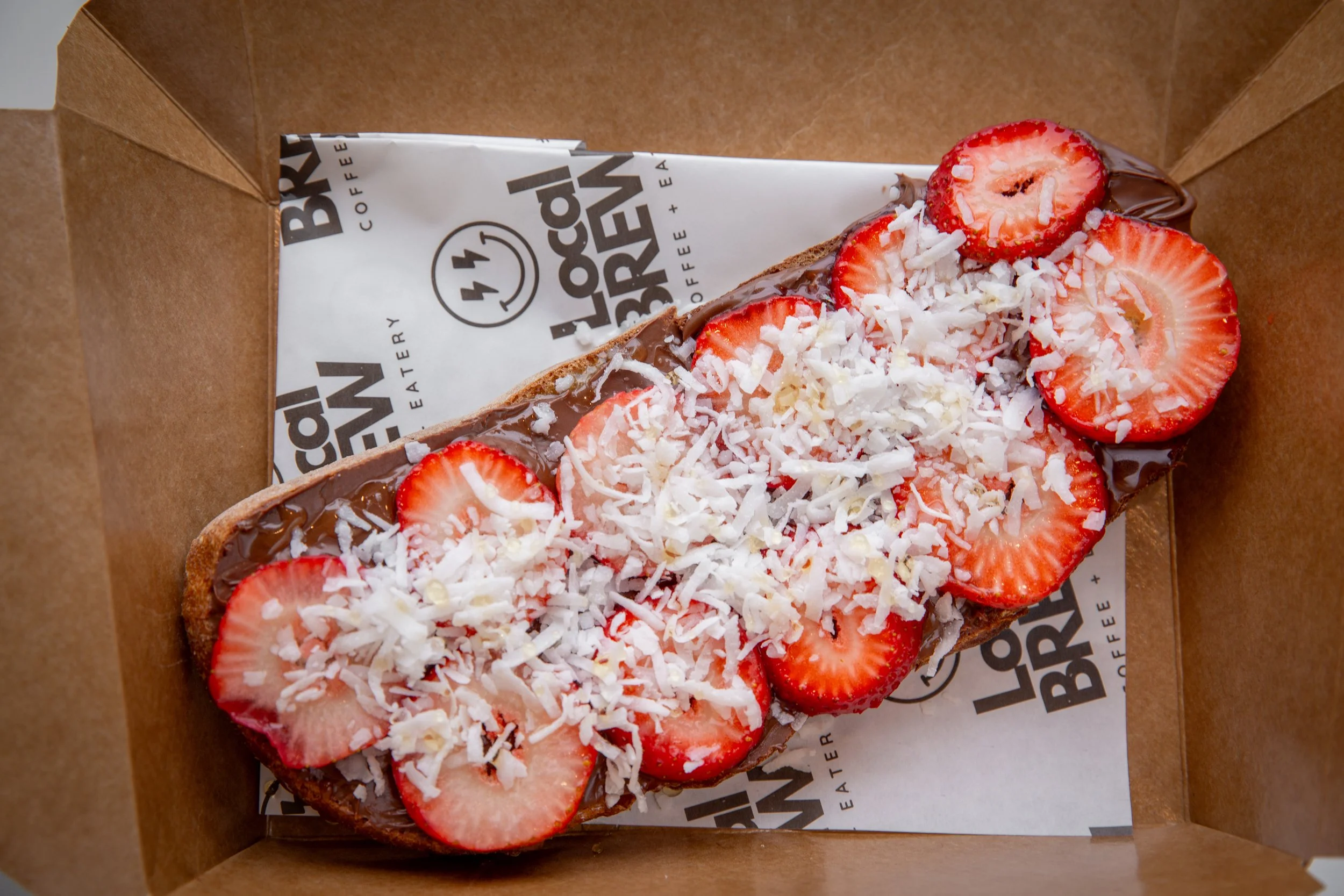 Chocolate-covered toast topped with sliced strawberries and shredded white chocolate inside a brown paper takeout box.