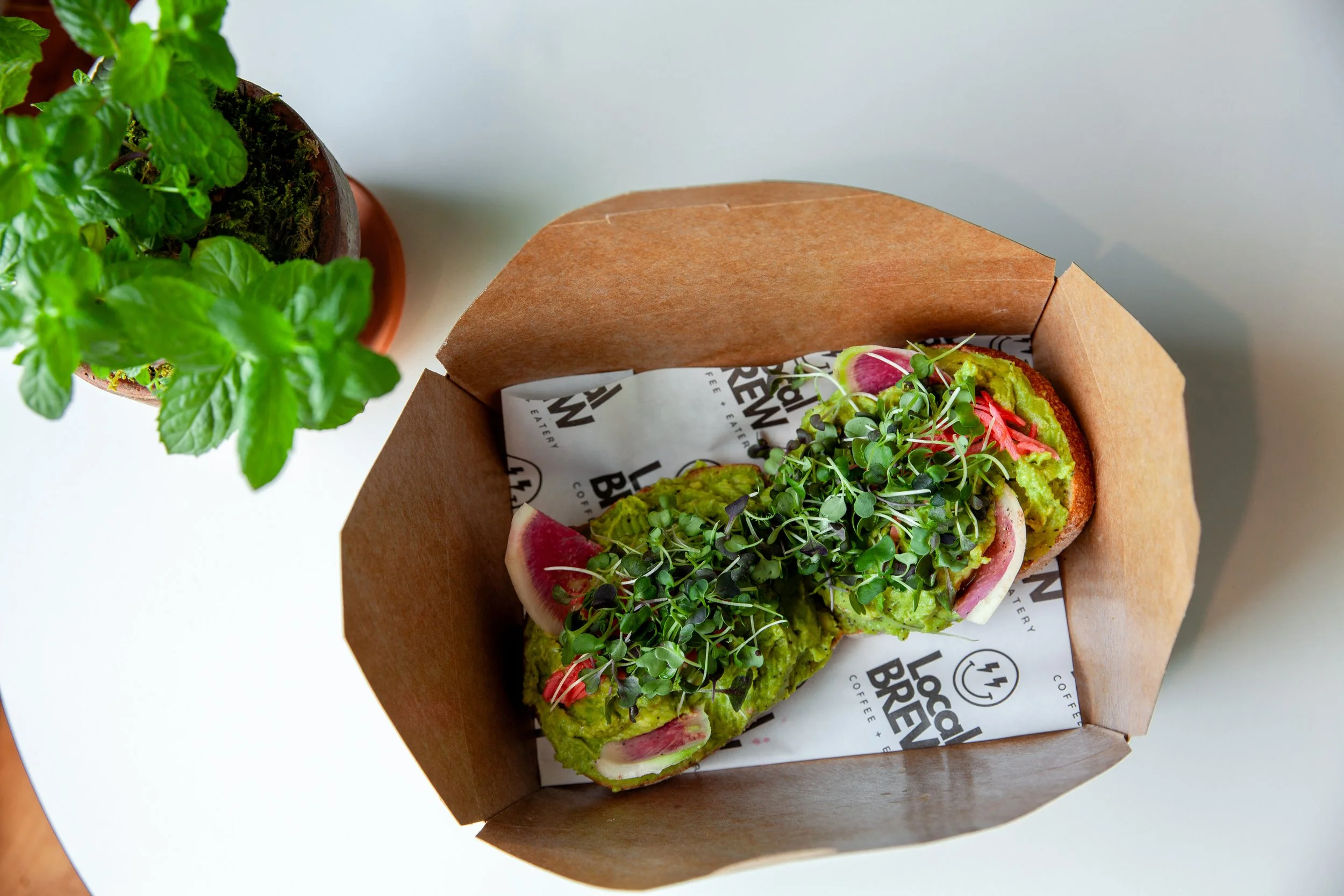 A cardboard tray of avocado toast topped with microgreens, radish slices, and mashed avocado, next to a potted mint plant on a white surface.
