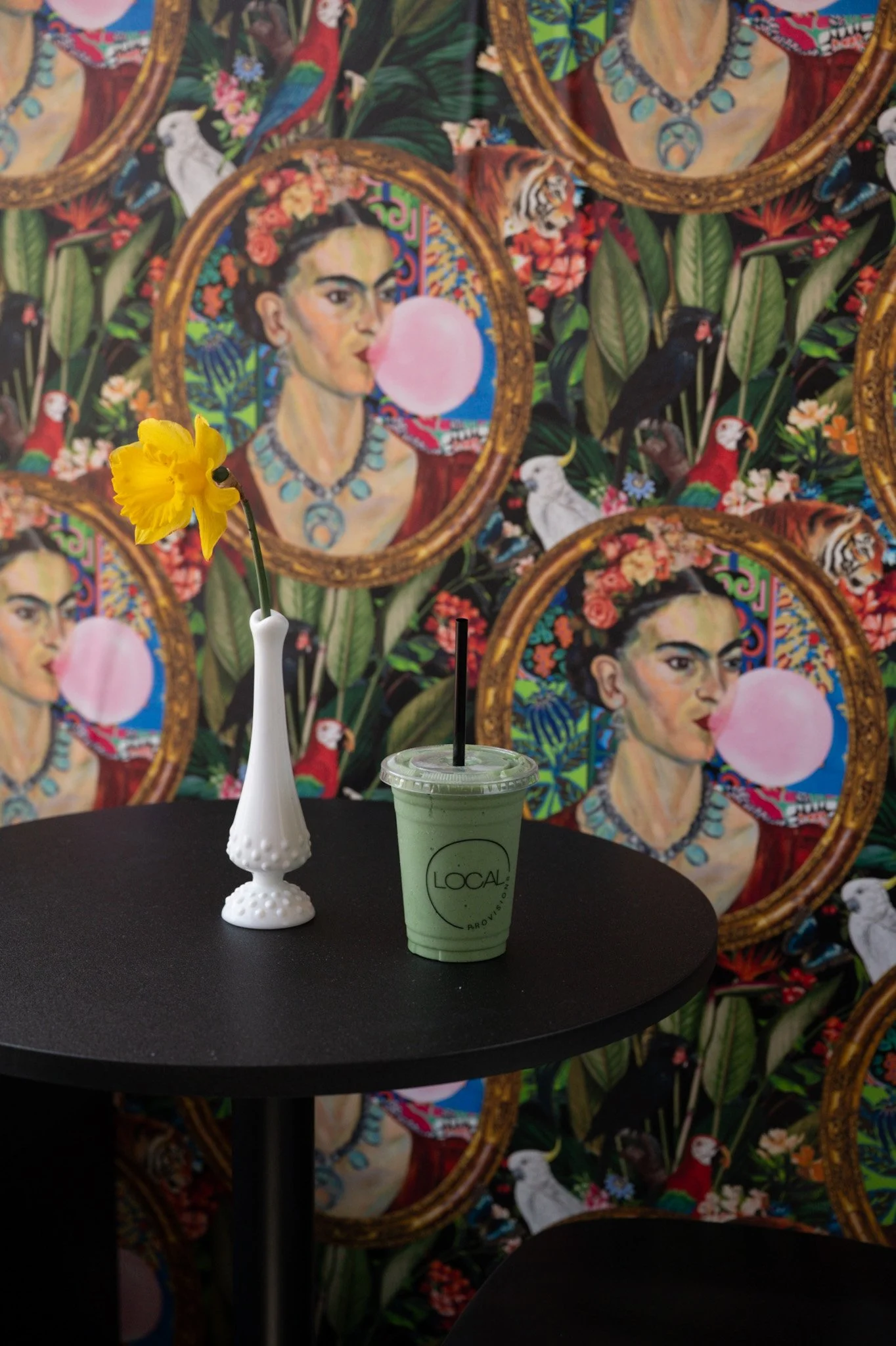 A small yellow flower in a tall white vase, a green smoothie drink with a straw on a black table, and a colorful background of artistic portraits and animals surrounded by leaves.