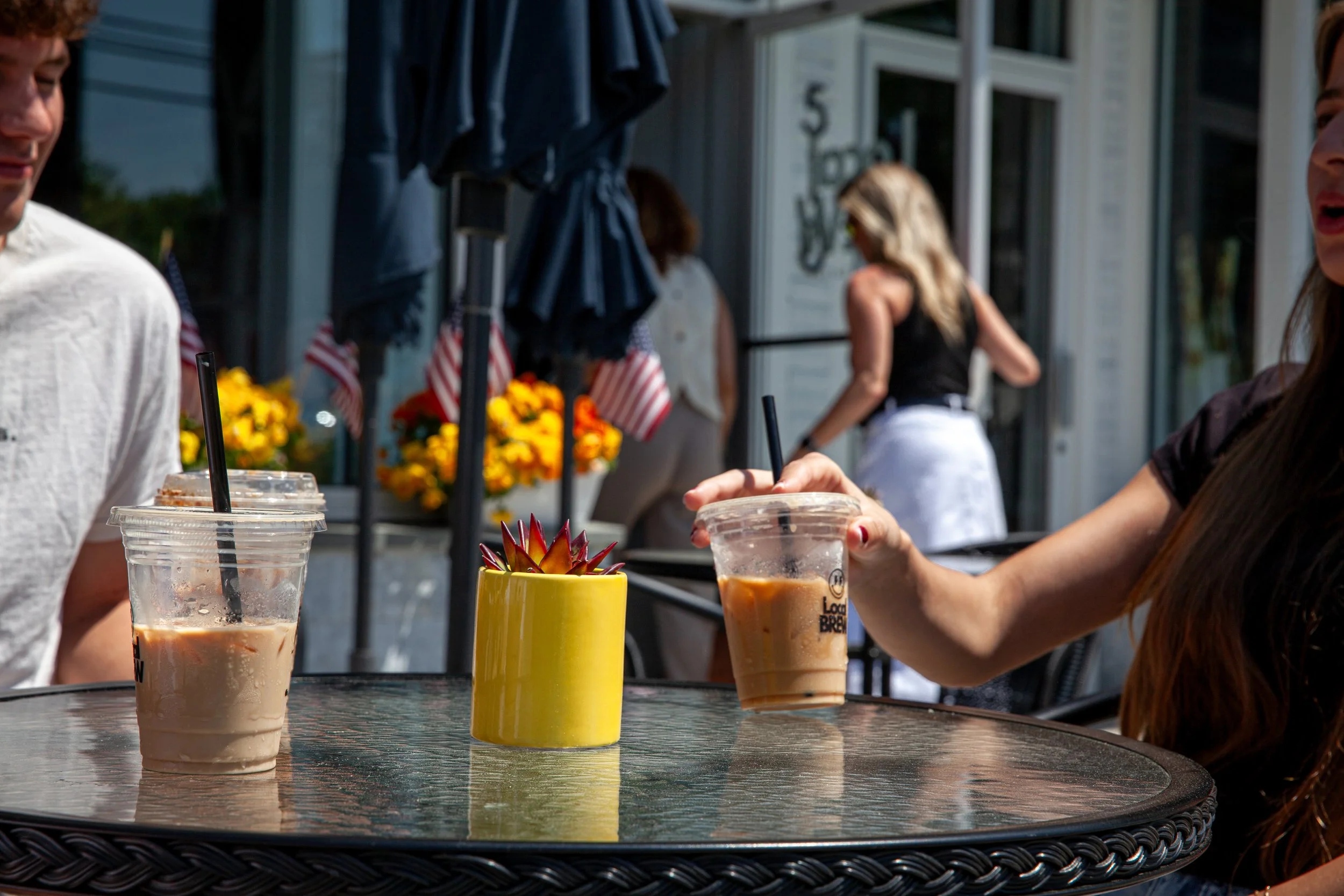 People sitting at an outdoor table with iced coffee drinks and a yellow container with a succulent plant, with people and a building in the background.
