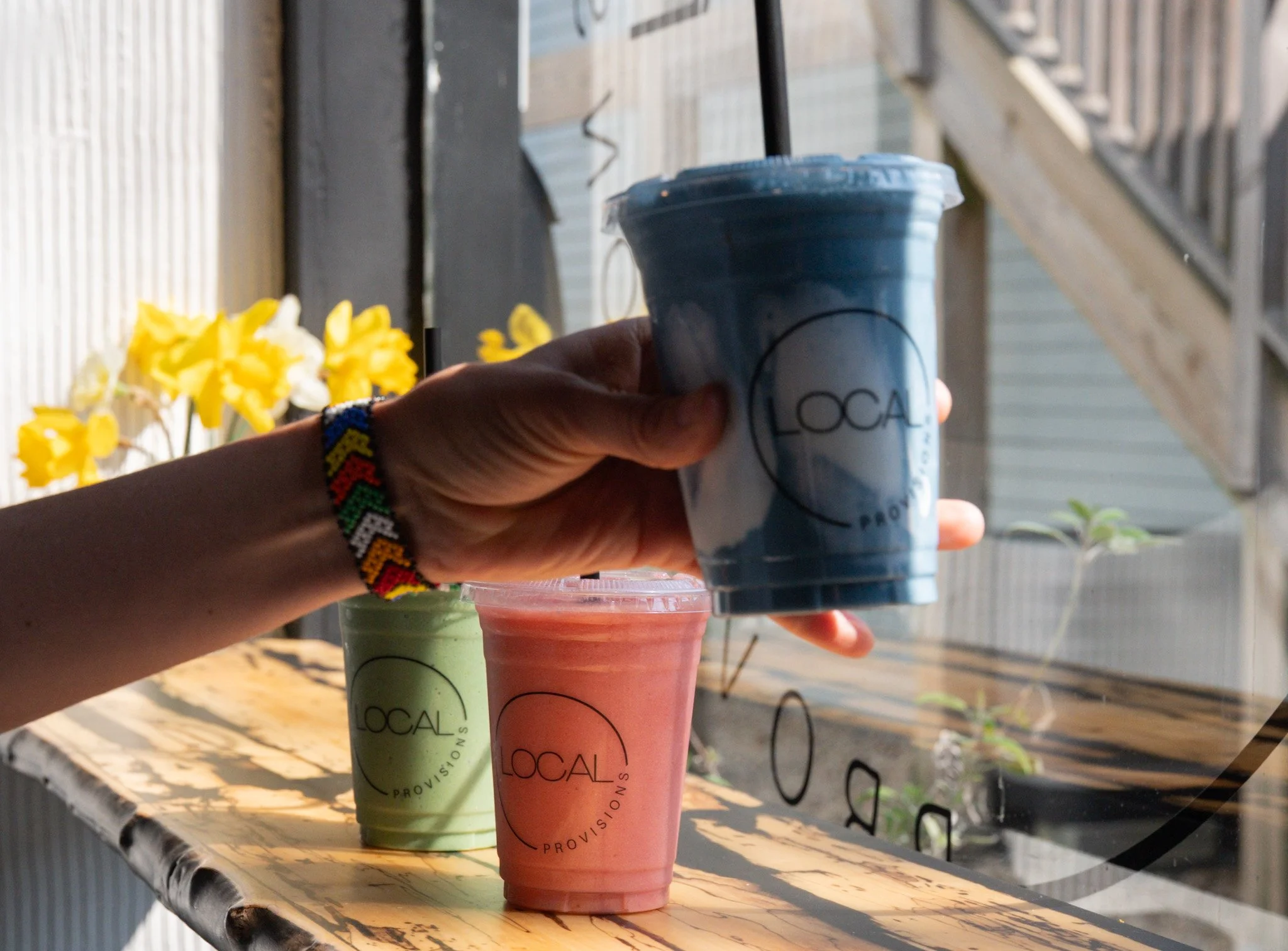 Hand holding three cups of smoothie in green, pink, and blue for beverage shop called 'Local Provisions' on a wooden table outdoors