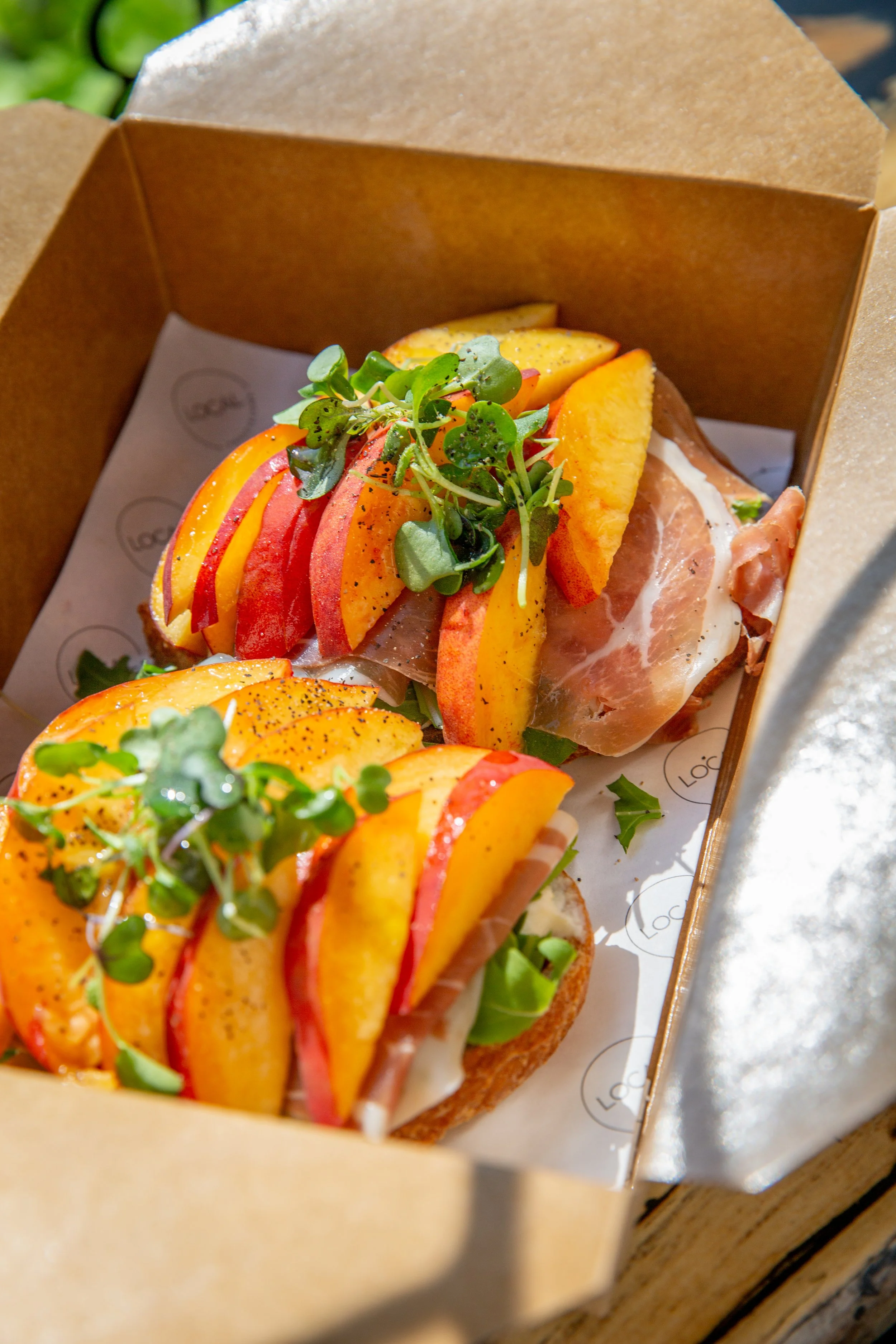 Open cardboard box containing three open-faced sandwiches topped with sliced peaches, sliced tomatoes, microgreens, and cracked black pepper.