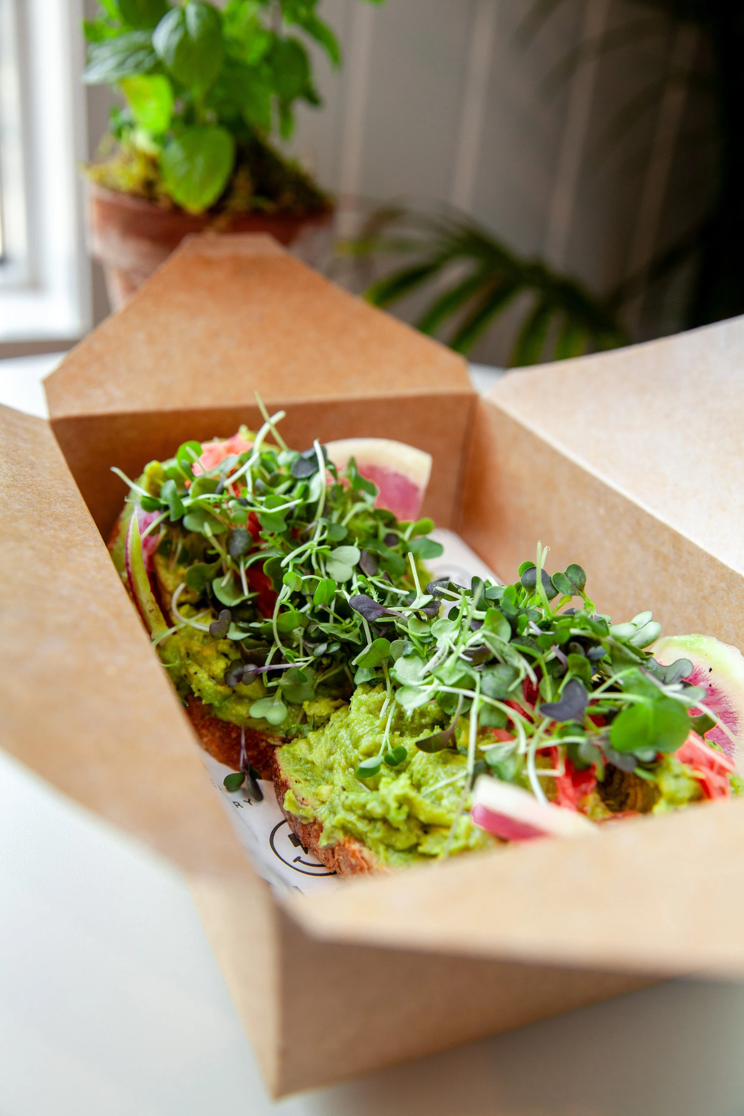Sliced avocado toast topped with microgreens, placed in a brown paper food container.