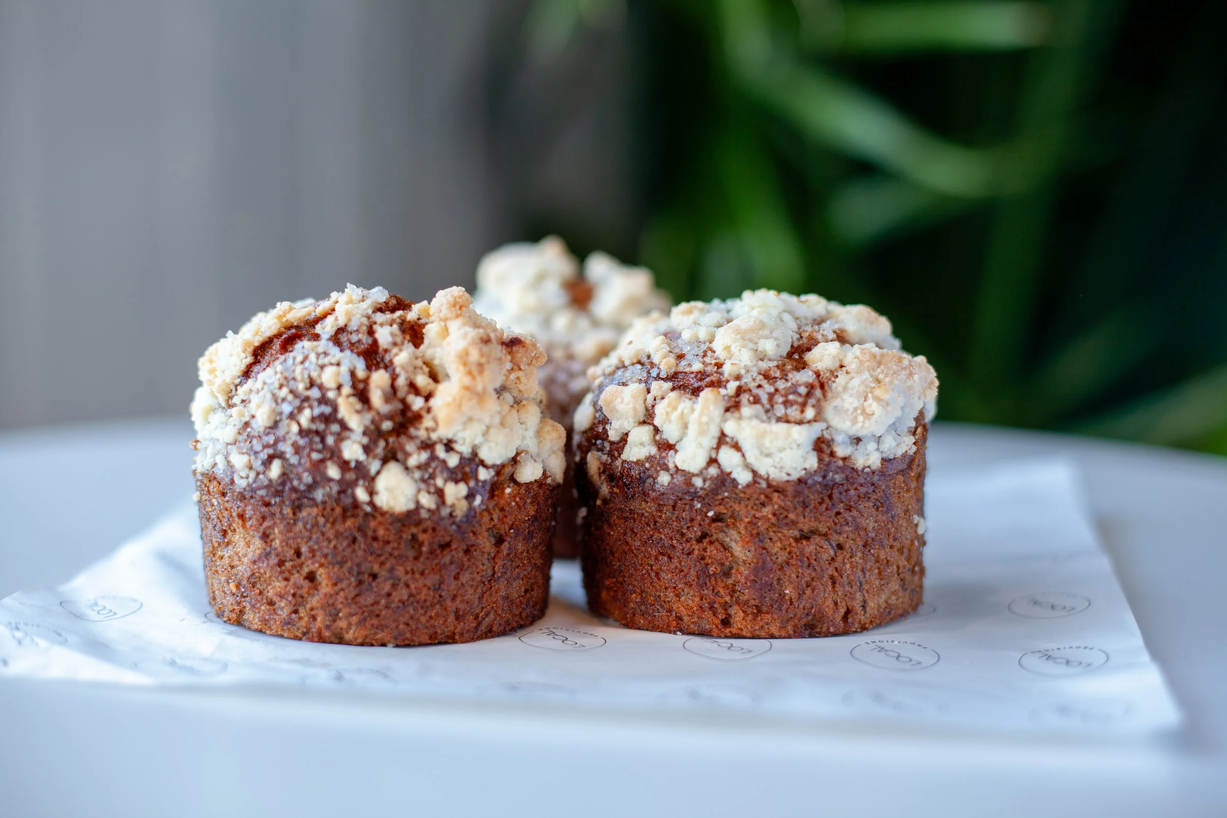 Two cruffins with a crumble topping sit on white paper with faint circular logos, against a blurred background of greenery.