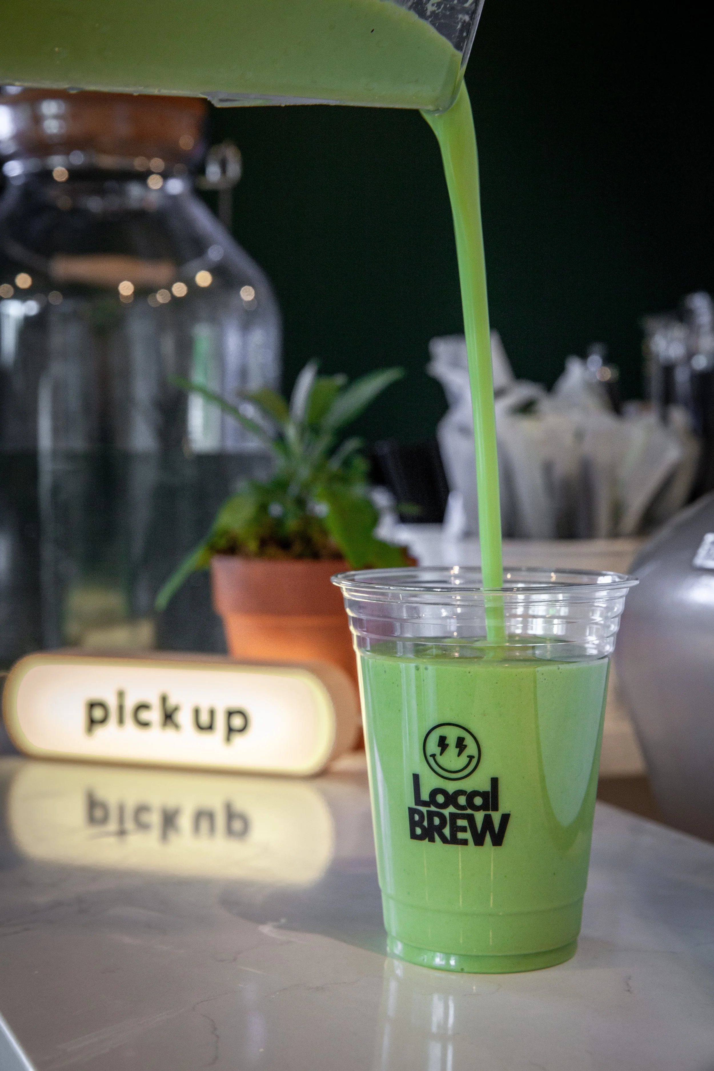 Green drink in a clear plastic cup with black text and smiley face logo, pouring from a container into a beverage cup, with a blurred background including a plant, a sign that says 'pickup' in different languages, and various objects.