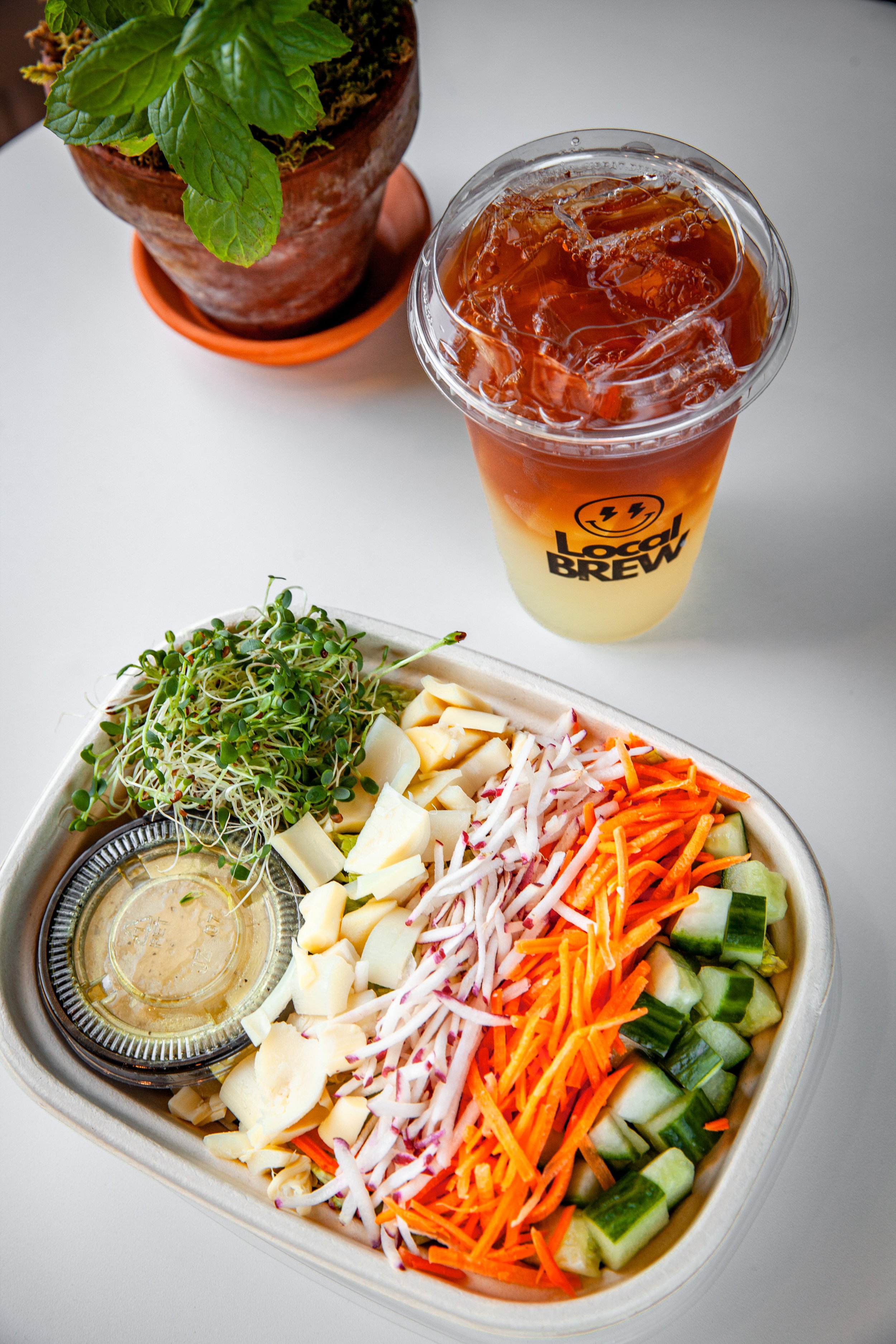 A salad with chopped vegetables including cucumbers, carrots, radishes, shredded cheese, and chopped boiled eggs on a metal platter, with a small container of dressing in the middle, placed on a white table. In the background, there is a potted plant and a beverage with ice in a cup labeled 'Local Brew.'