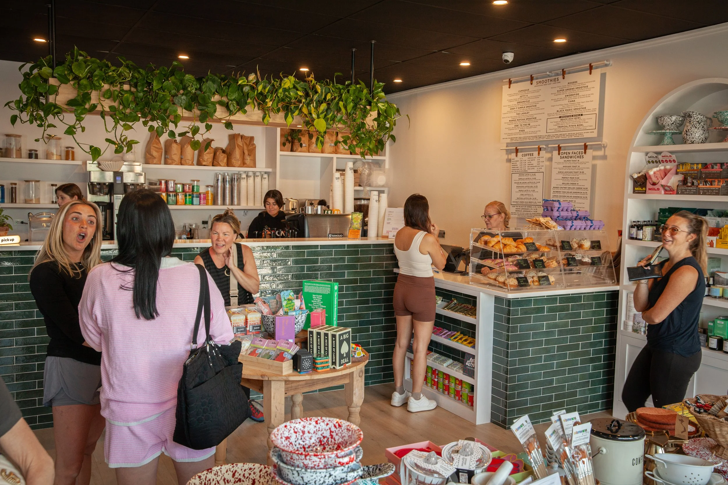 Inside a cozy cafe with customers ordering and chatting. The counter displays pastries and baked goods, with a menu hanging on the wall. Green plants hang from the ceiling, and shelves are stocked with coffee supplies and snacks.