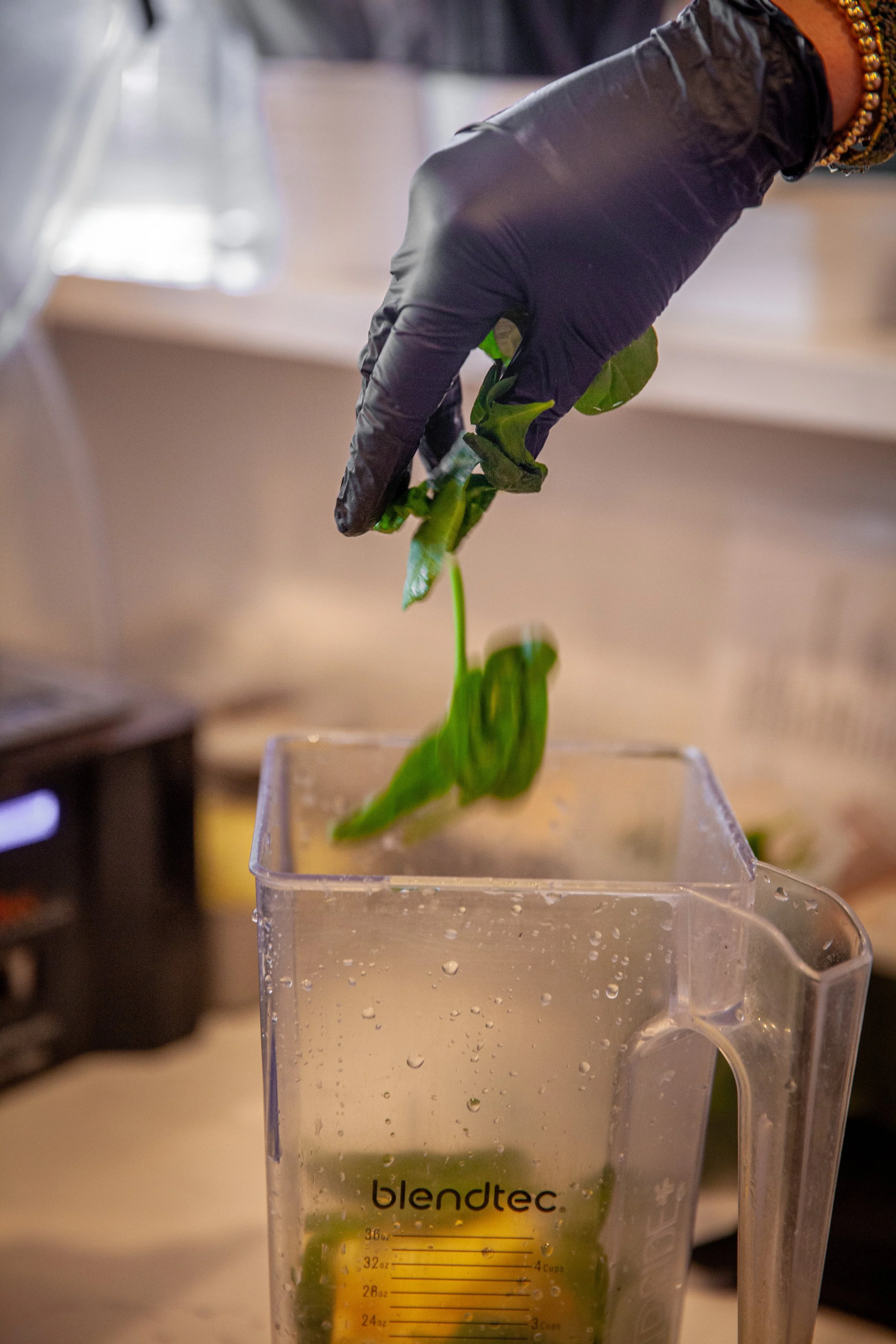 Close-up of a hand wearing a black glove holding a green leaf with a stem over a clear plastic measuring cup filled with a green liquid. The measuring cup has the brand name 'Blendtec' and measurement markings visible, placed on a countertop.