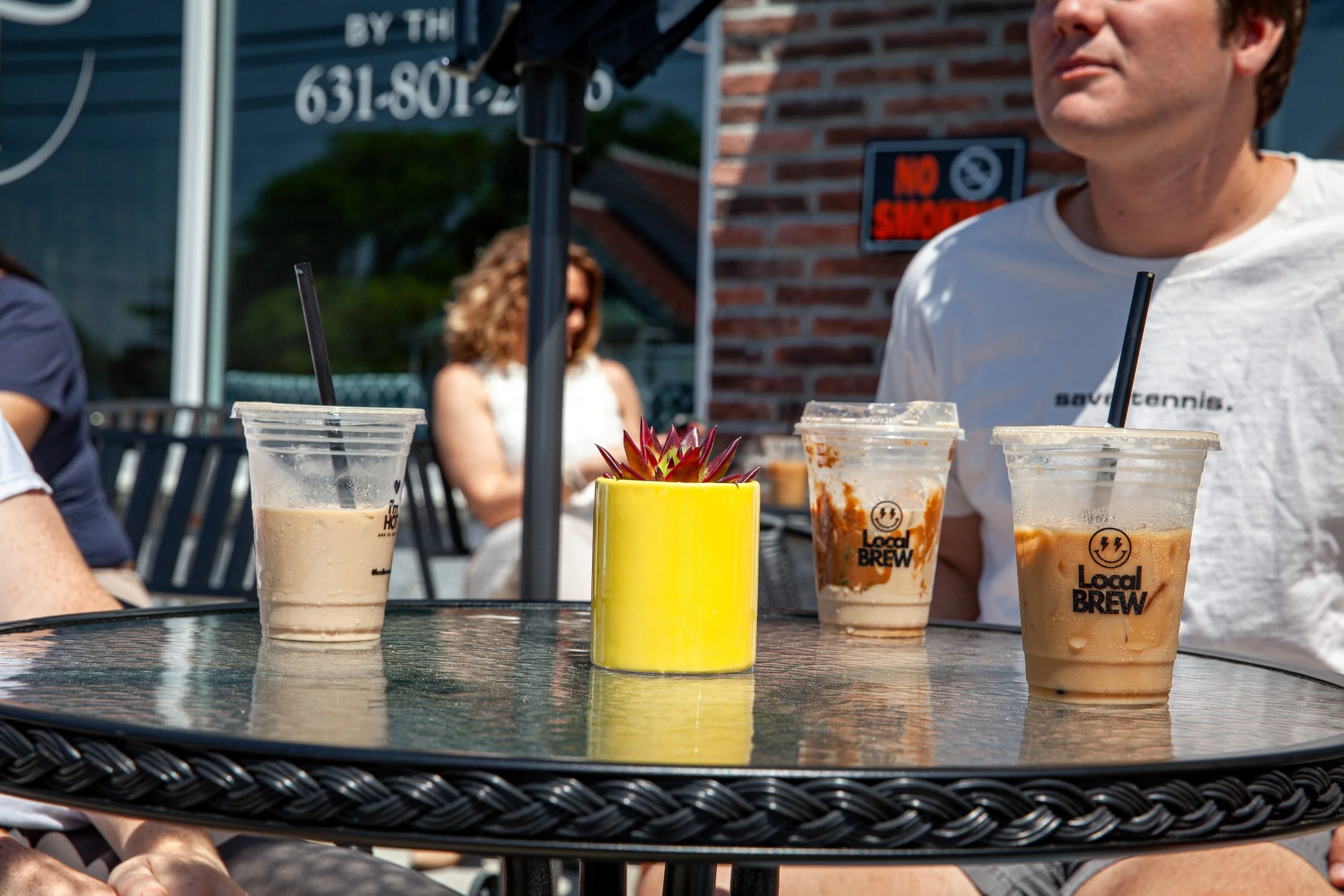 Three iced coffee drinks on a black wicker outdoor table, with a small yellow pot containing a succulent plant and a black straw in each cup. People are sitting in the background at a casual outdoor setting.