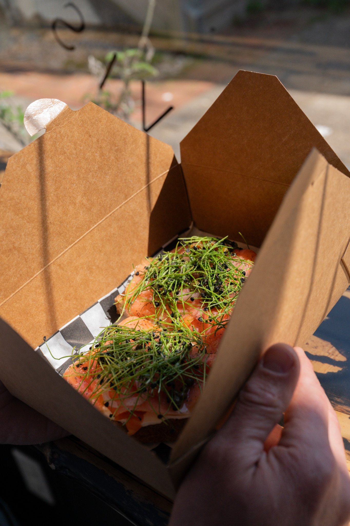 A person holding a cardboard takeout container with smoked salmon topped with microgreens and black caviar.