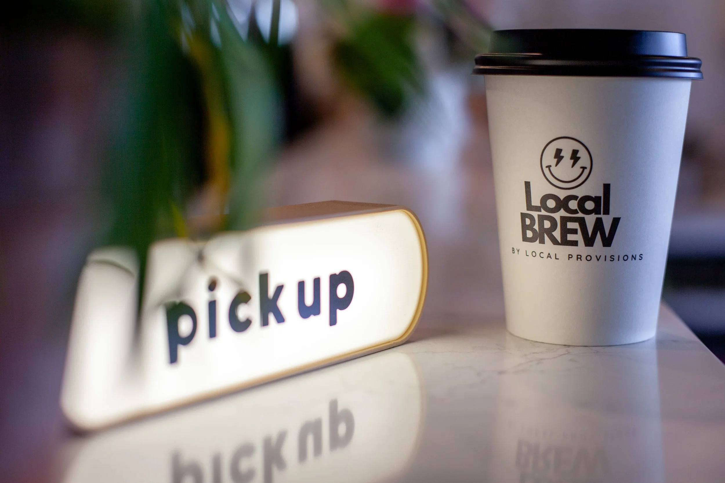 A white disposable coffee cup with a black lid, displaying a smiling face with lightning bolt eyes and the text "Local BREW" underneath, placed on a table near a sign that says "pickup" in colorful letters, with some blurred greenery in the background.