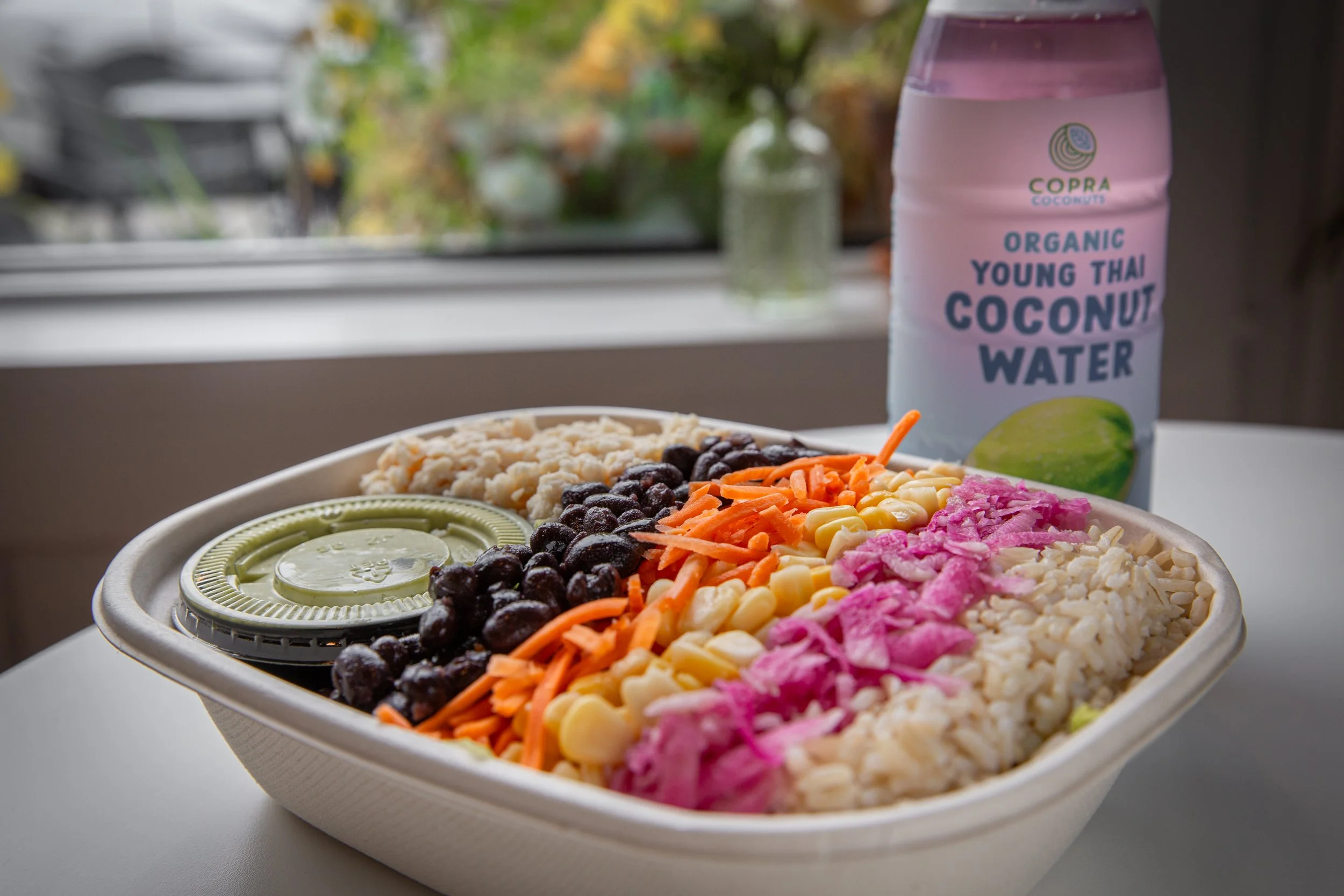 A takeout bowl containing a variety of colorful shredded vegetables, black beans, and cheese, with slices of cucumber and a container of lime, and a carton of organic young Thai coconut water in the background.