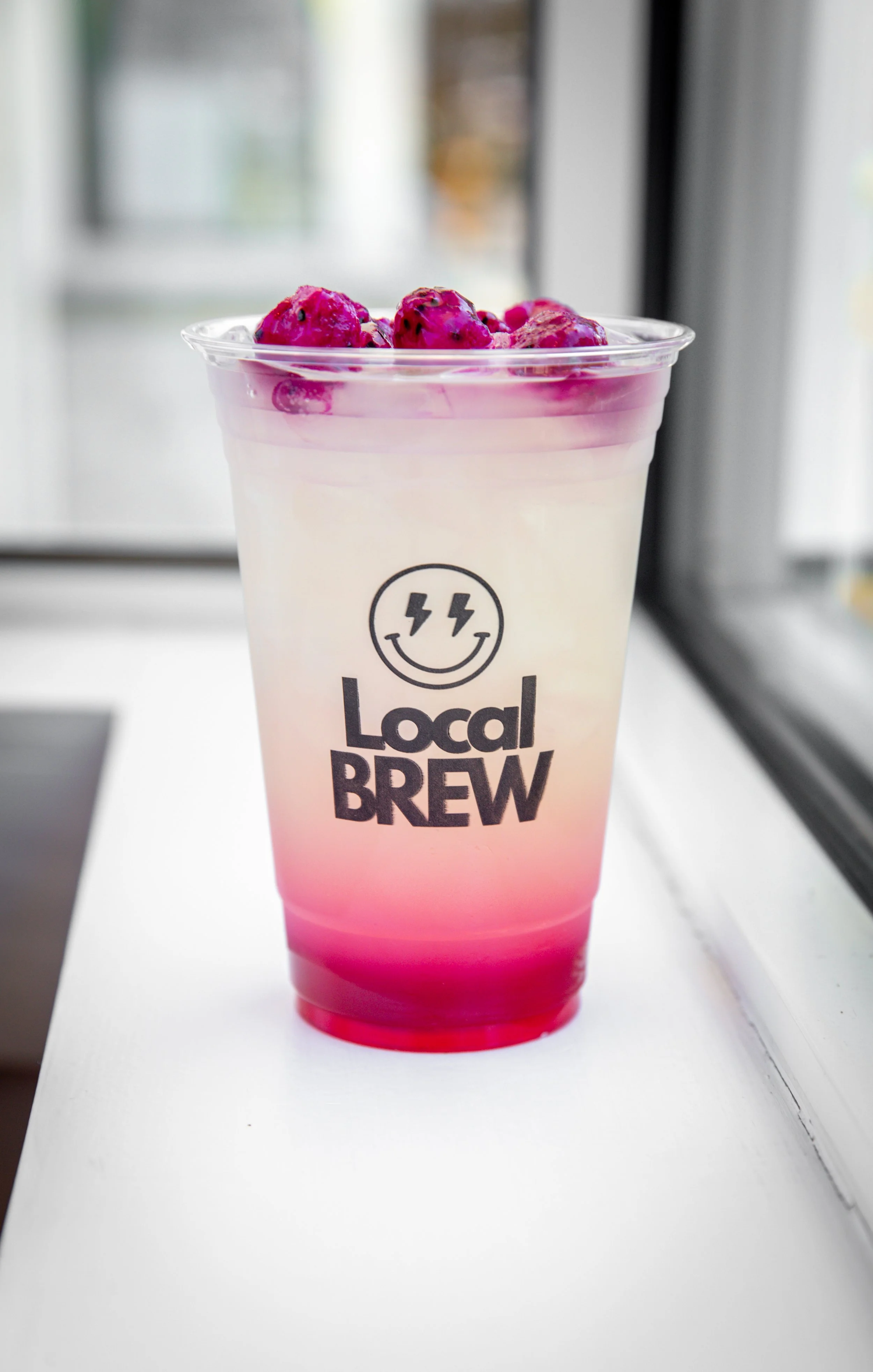A clear plastic cup with a pink gradient at the bottom, filled with a white beverage topped with pink berries, and the logo 'Local Brew' with a smiling face and lightning bolts in the center.
