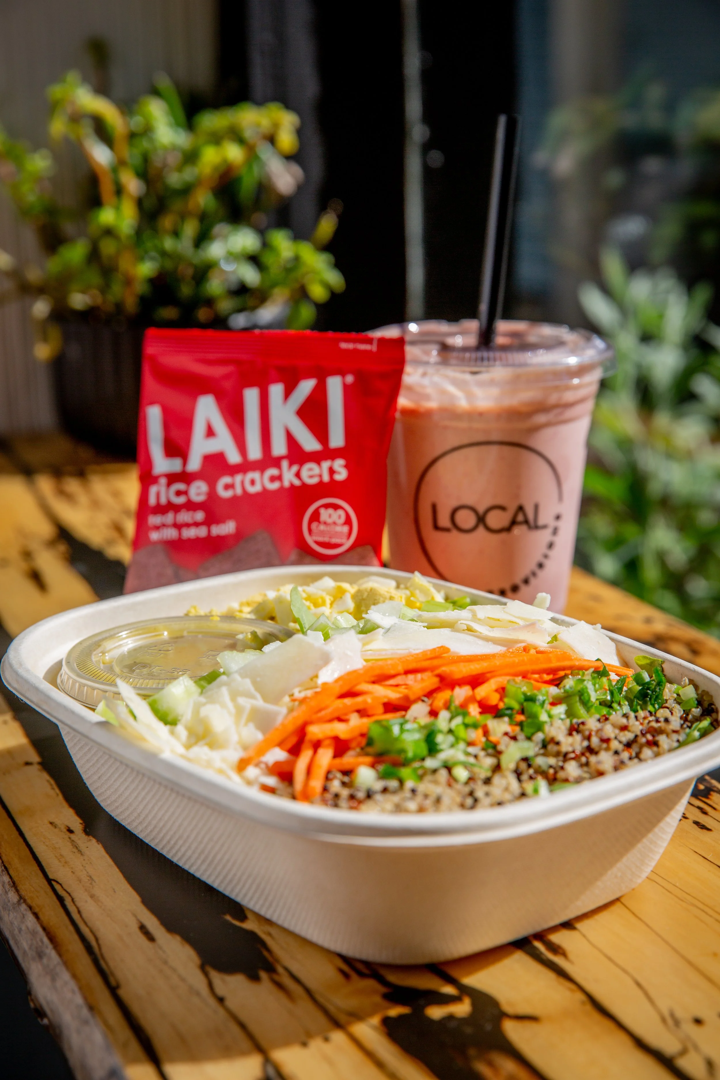A takeout container filled with rice, chopped vegetables, and shredded cheese, with a container of sauce on top, on a wooden table. In the background, a bag of Laiki rice crackers, a pink drink with black straws, and a potted plant.