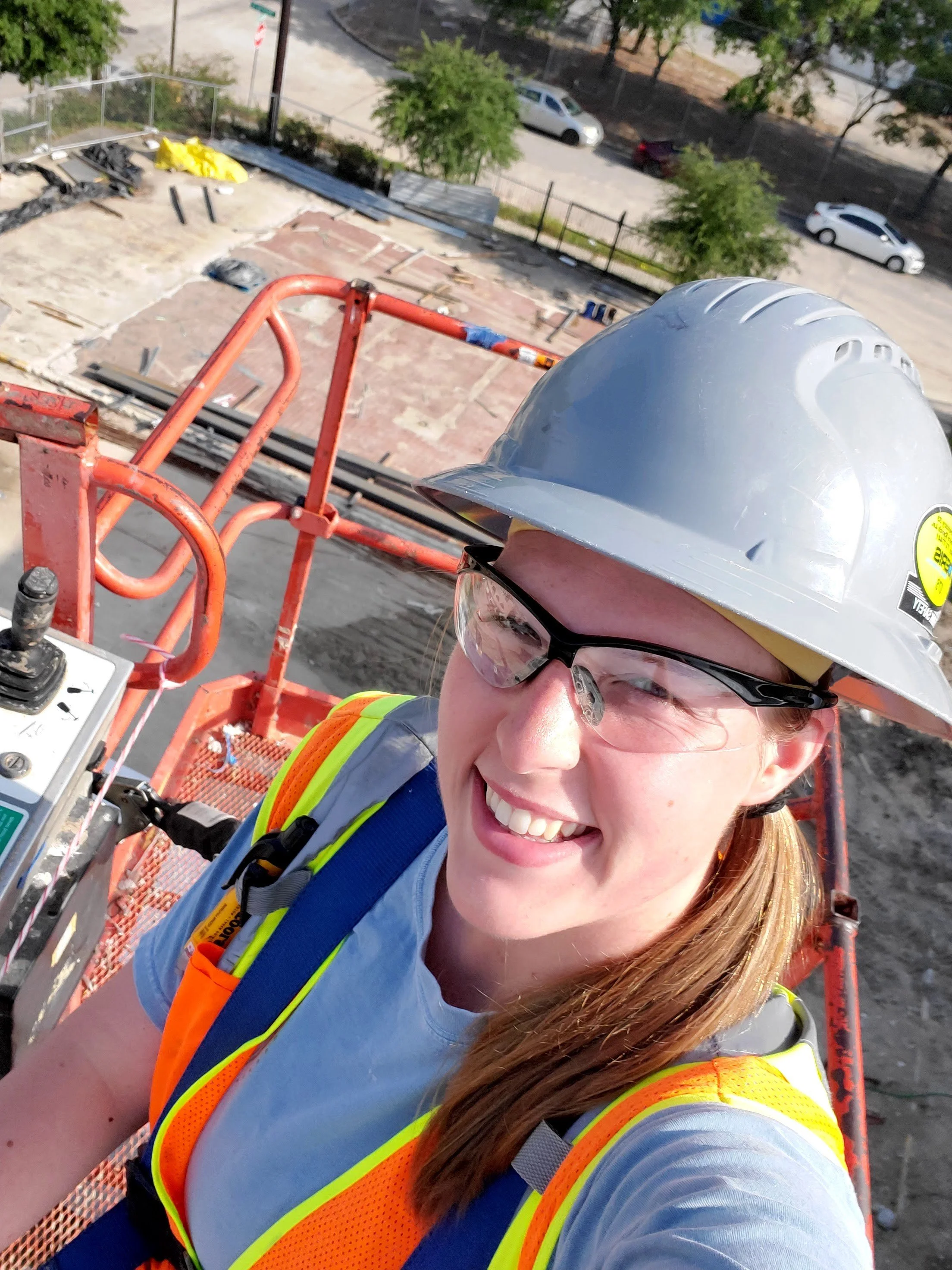A person wearing a hard hat and safety glasses smiling while taking a selfie from a construction lift platform.