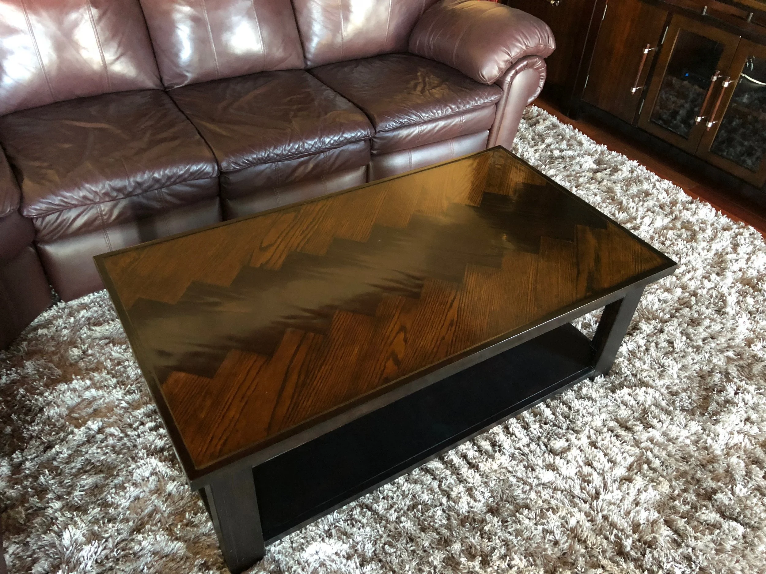 A living room with a brown leather sofa, a wooden coffee table with a zigzagged pattern, a white shaggy rug, and a dark wooden cabinet with glass doors.