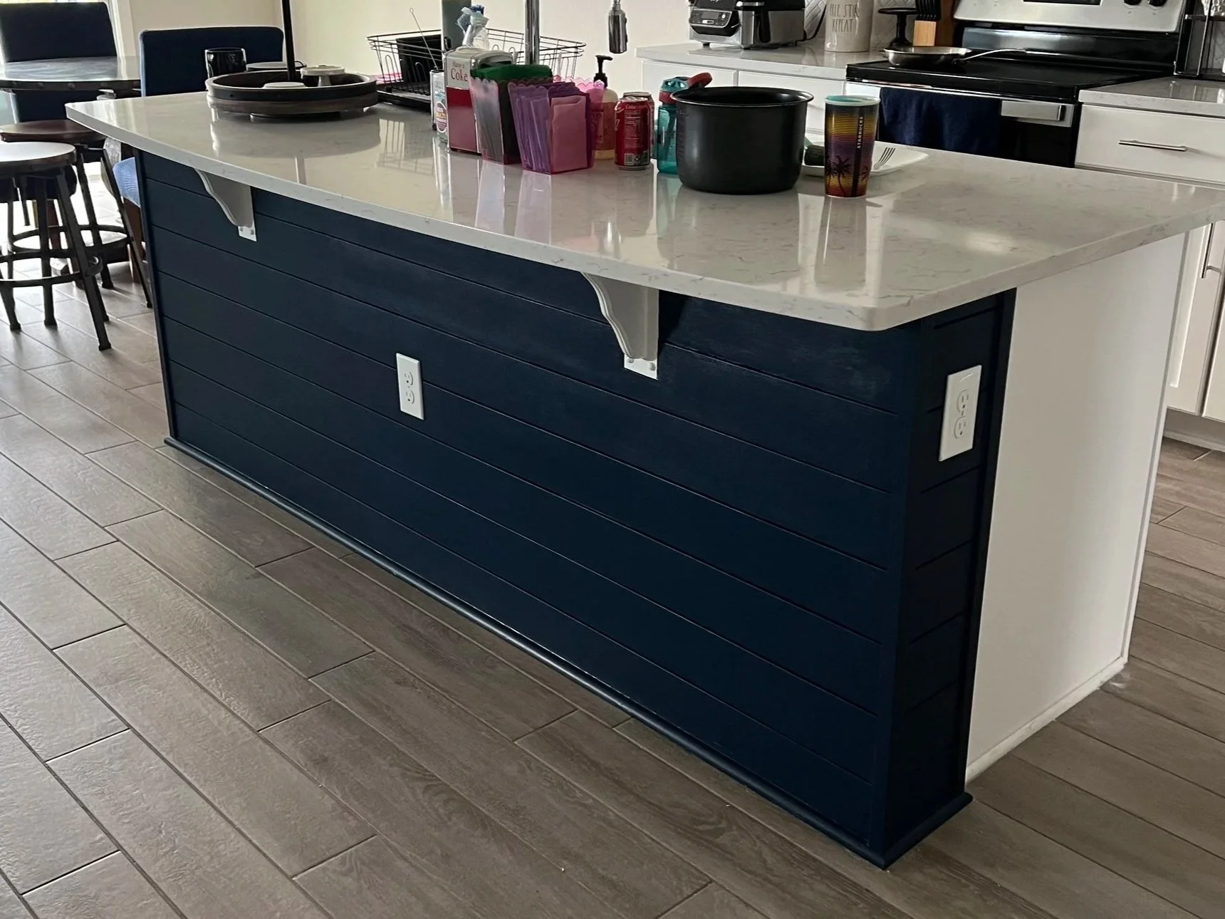 Kitchen island with a white marble top and blue paneled sides, with electrical outlets on the side. Items on top include bowls, a black pot, a glass, condiments, and a variety of colorful items. The background shows a white countertop, stove, coffee 