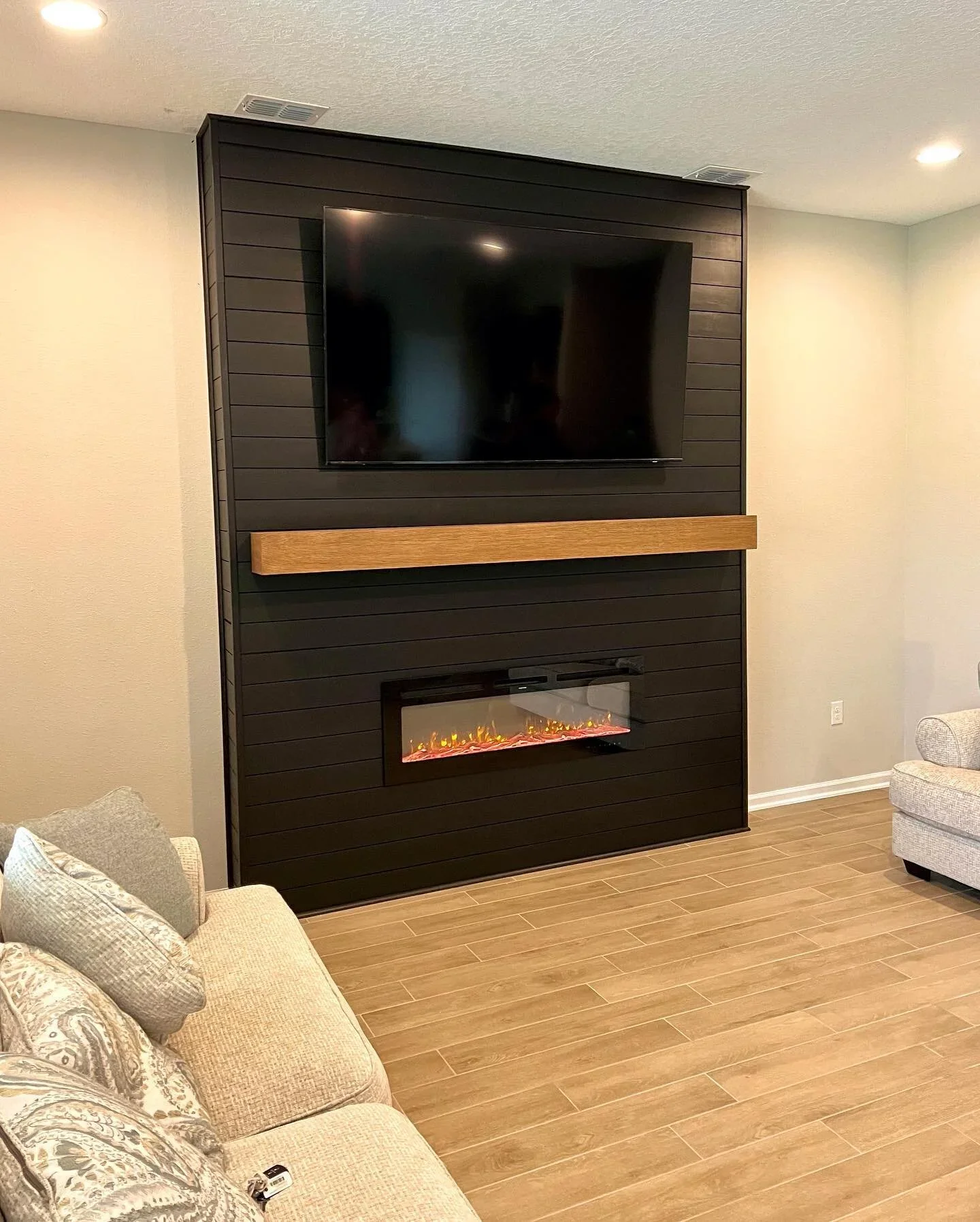 Living room with beige sofa, patterned cushions, small beige armchair, wood-look floor, black wall with mounted TV, wooden mantel, and electric fireplace below the TV.