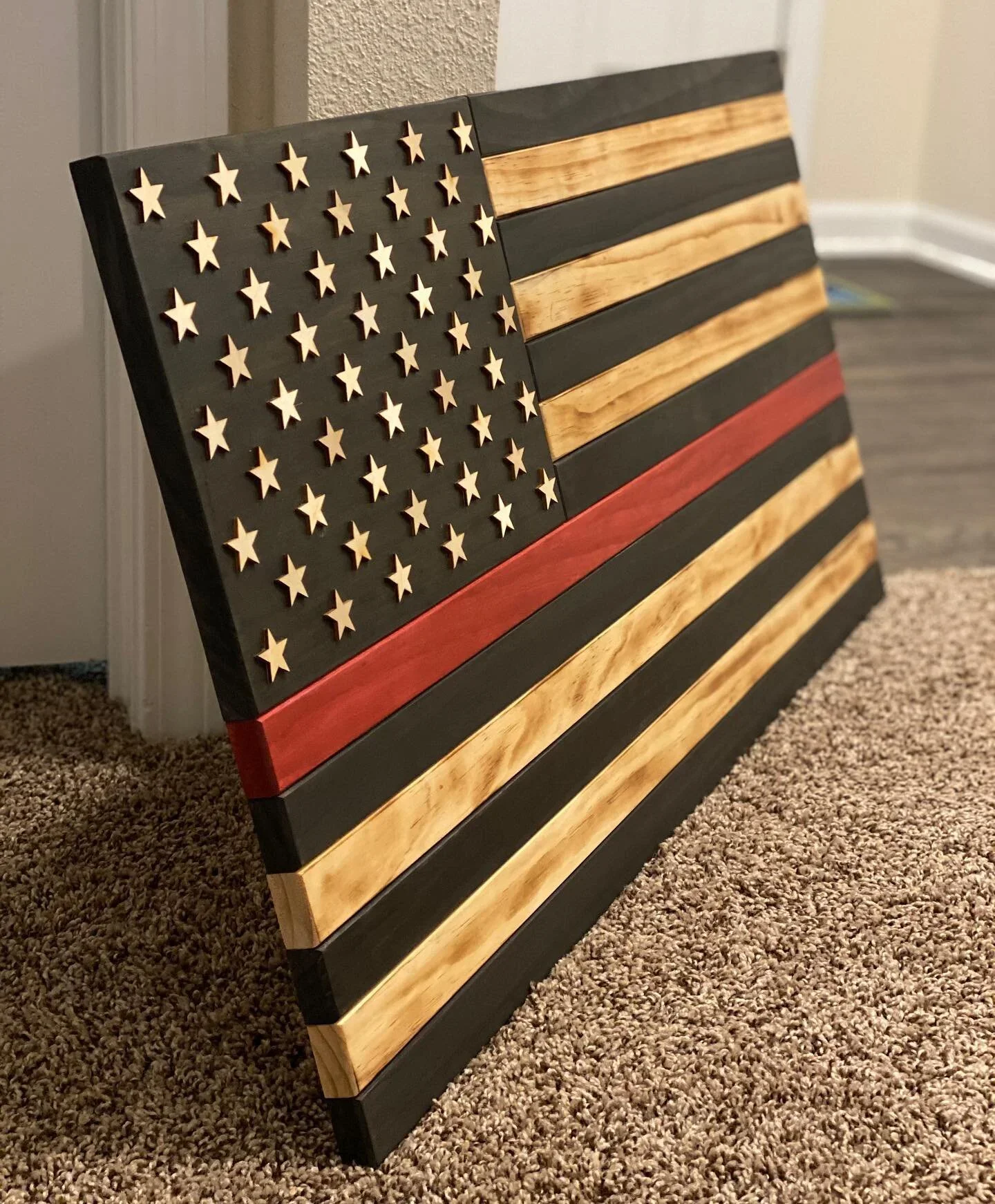 A horizontal American flag-themed wooden wall art piece with black, natural wood, and red stripes, and a black field with wooden stars in place of stars.