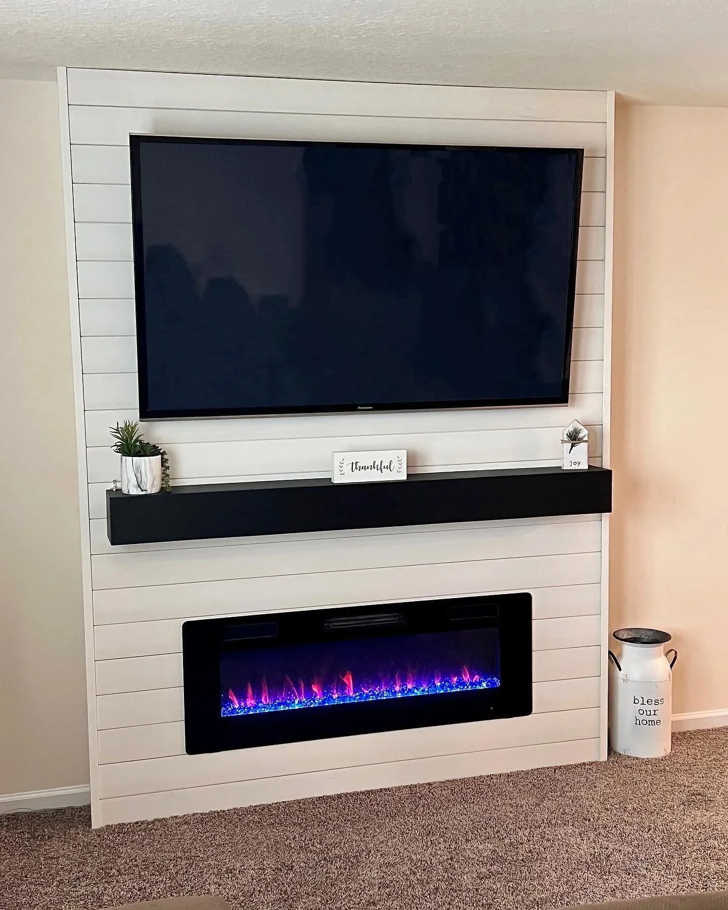 Living room corner with white shiplap wall, a flat-screen TV, a linear electric fireplace below, and decorative items including potted plants, a small sign reading 'thankful', and a container with a plant and the words 'bless our home' on it.