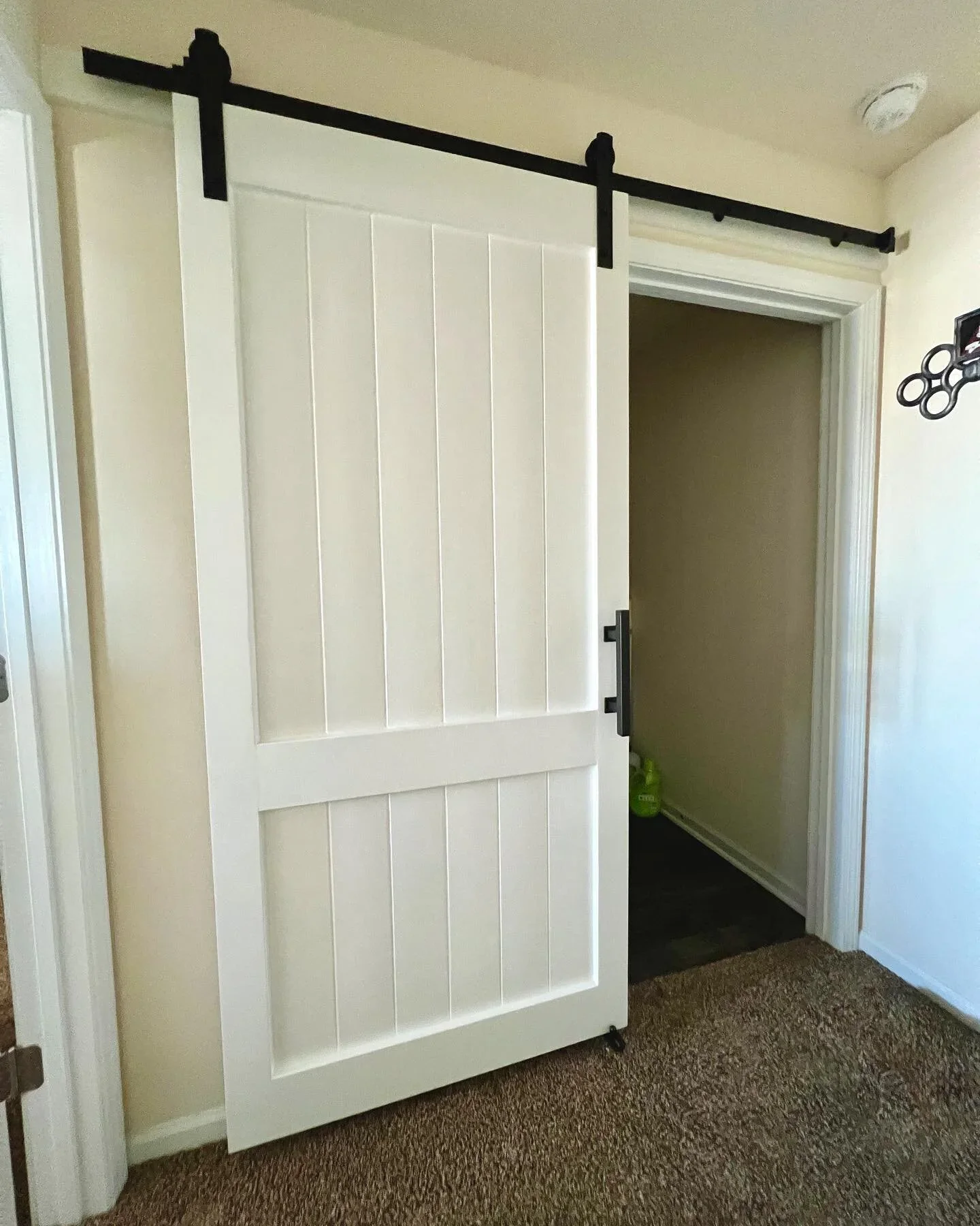 A white sliding barn door with black hardware, partially open, revealing a small entryway with a green bottle on the floor.