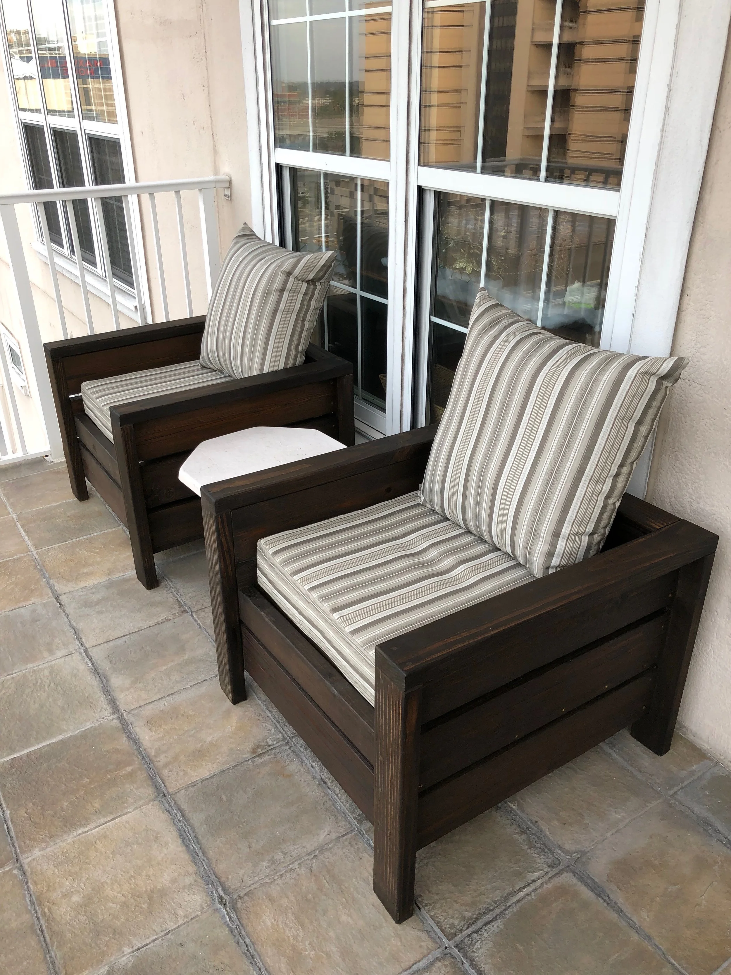 Two wooden chairs with striped cushions on a balcony with a sliding glass door and a railing.