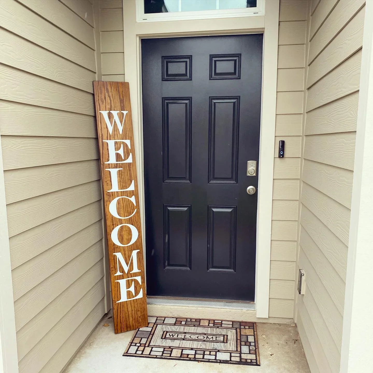 Front porch with a black door, a welcome mat, a wooden 'Welcome' sign, and beige vinyl siding.